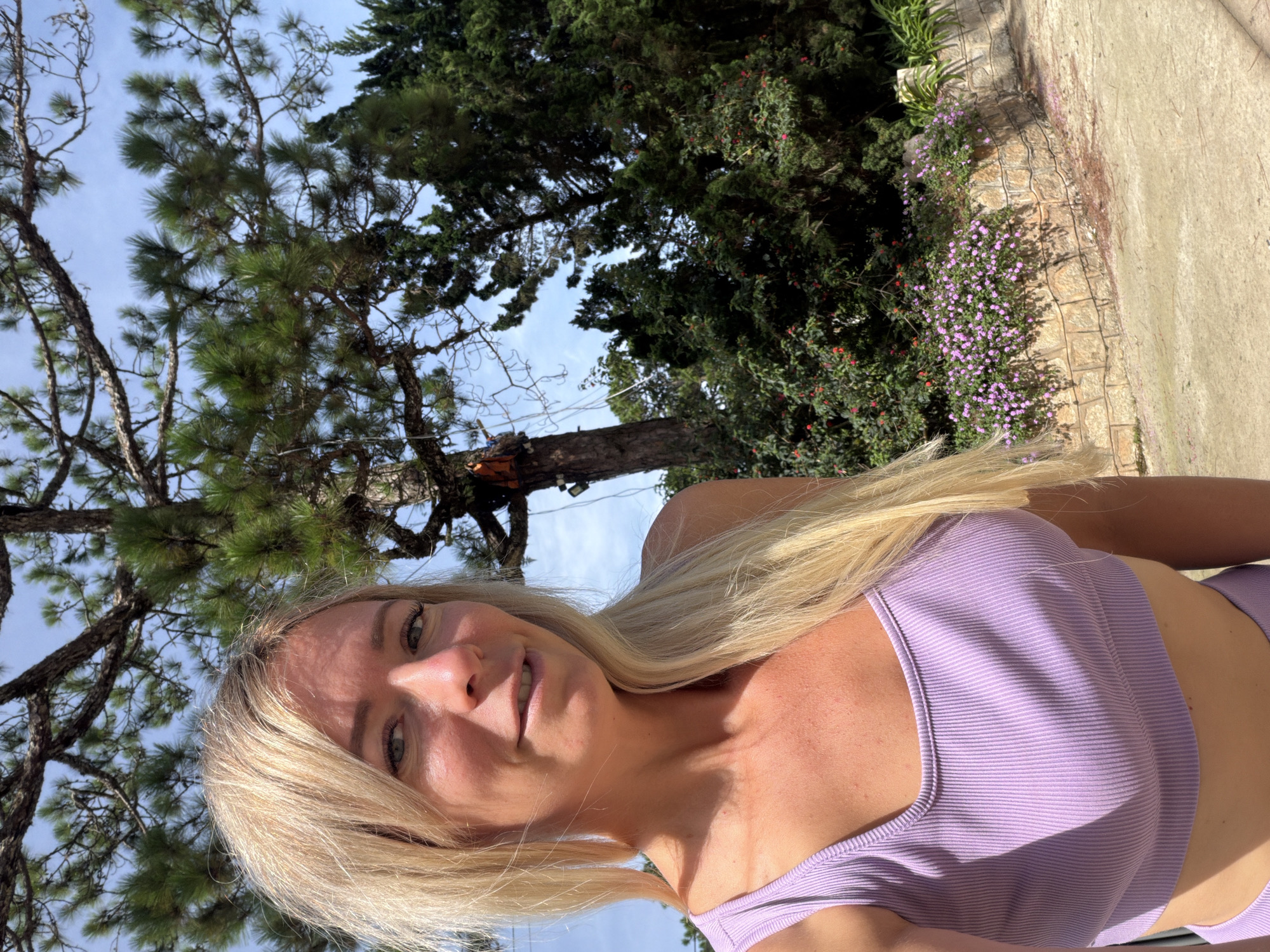 A blonde woman with shoulder-length hair is taking a selfie outdoors. She is wearing a light purple athletic top. Behind her, a large pine tree with dark green needles is visible, along with a clear blue sky and lush green foliage. In the background, there's a stone wall with small purple flowers growing on top. The scene appears to be in a park or garden setting in Xuân Trường, Đà Lạt, Vietnam.