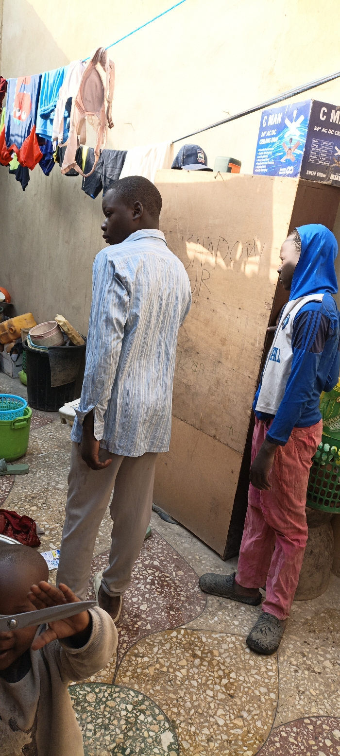 The image portrays an outdoor domestic scene in Kaura Goje, Nigeria. Three individuals are present on a multi-colored terrazzo-style tiled floor. In the foreground, a child, partially cropped, holds a pair of silver-handled scissors. Standing center-left, a male in a light blue and white striped long-sleeve shirt and light brown trousers faces right, away from the camera. To his right, a younger male, seen in profile, wears a blue hooded long-sleeve shirt with white and blue chest detailing, pink trousers, and dark slip-on shoes, also facing right.
Behind the two standing males is a large, vertical light-brown wooden panel bearing dark, handwritten graffiti, including "SNAROWY JR". To the left, various containers are stacked, including a black bucket, a white bucket, and yellow jerrycans. Above, a blue rope clothesline extends across the upper frame, draped with assorted garments, including t-shirts and a light-colored bra. In the upper right, a "C MAN 24" AC/DC CEILING FAN" cardboard box is visible on a surface, next to a dark blue baseball cap. The background features light-colored walls, and the scene is illuminated by bright daylight.