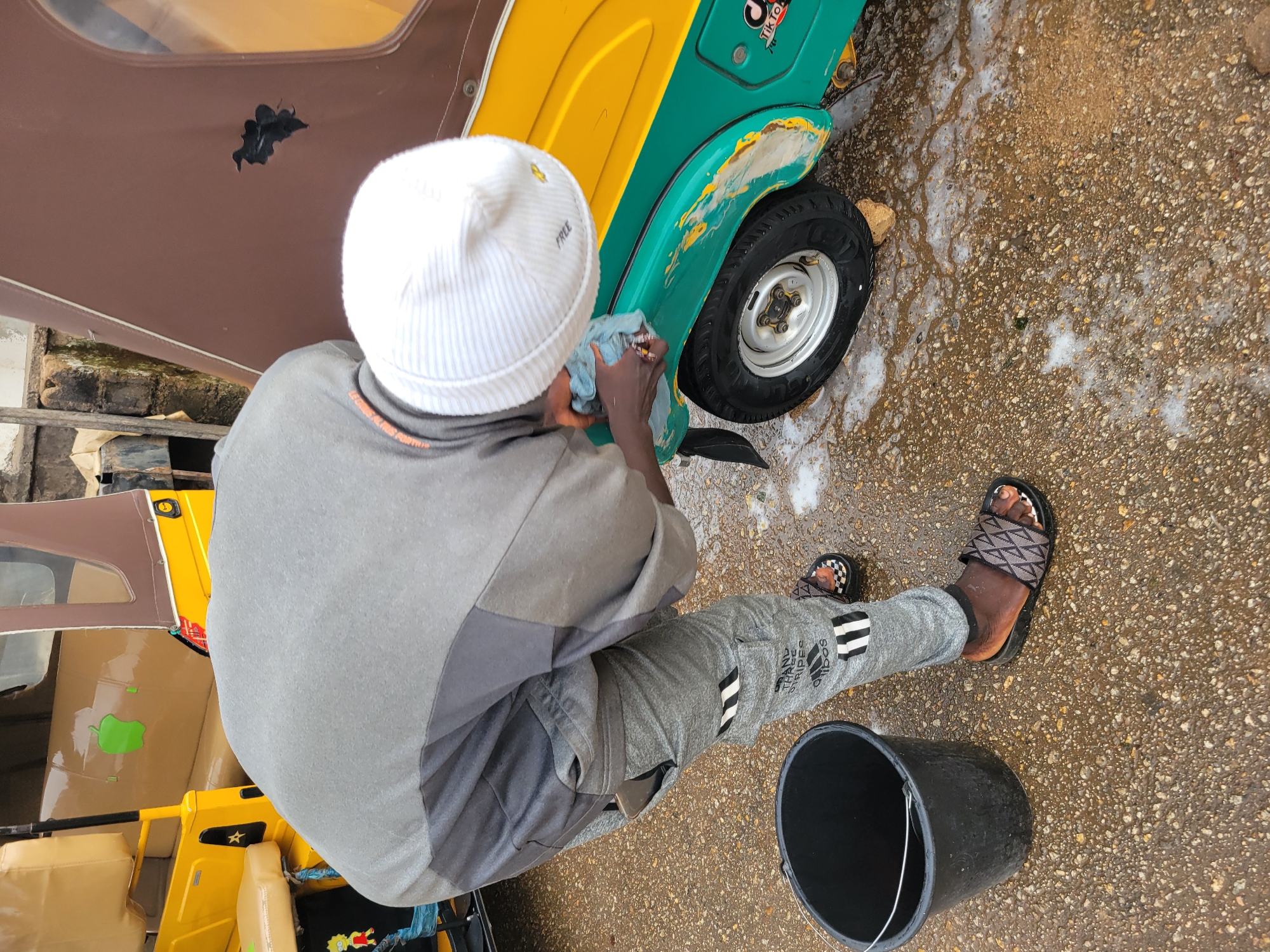 A man is washing an auto-rickshaw in Jos, Nigeria. The man is wearing a white beanie, a grey t-shirt, grey sweatpants with Adidas stripes, and sandals. He is holding a blue cloth and cleaning the front wheel well of the green and yellow auto-rickshaw. A black bucket sits next to his feet on the wet pavement. Other auto-rickshaws are parked nearby.