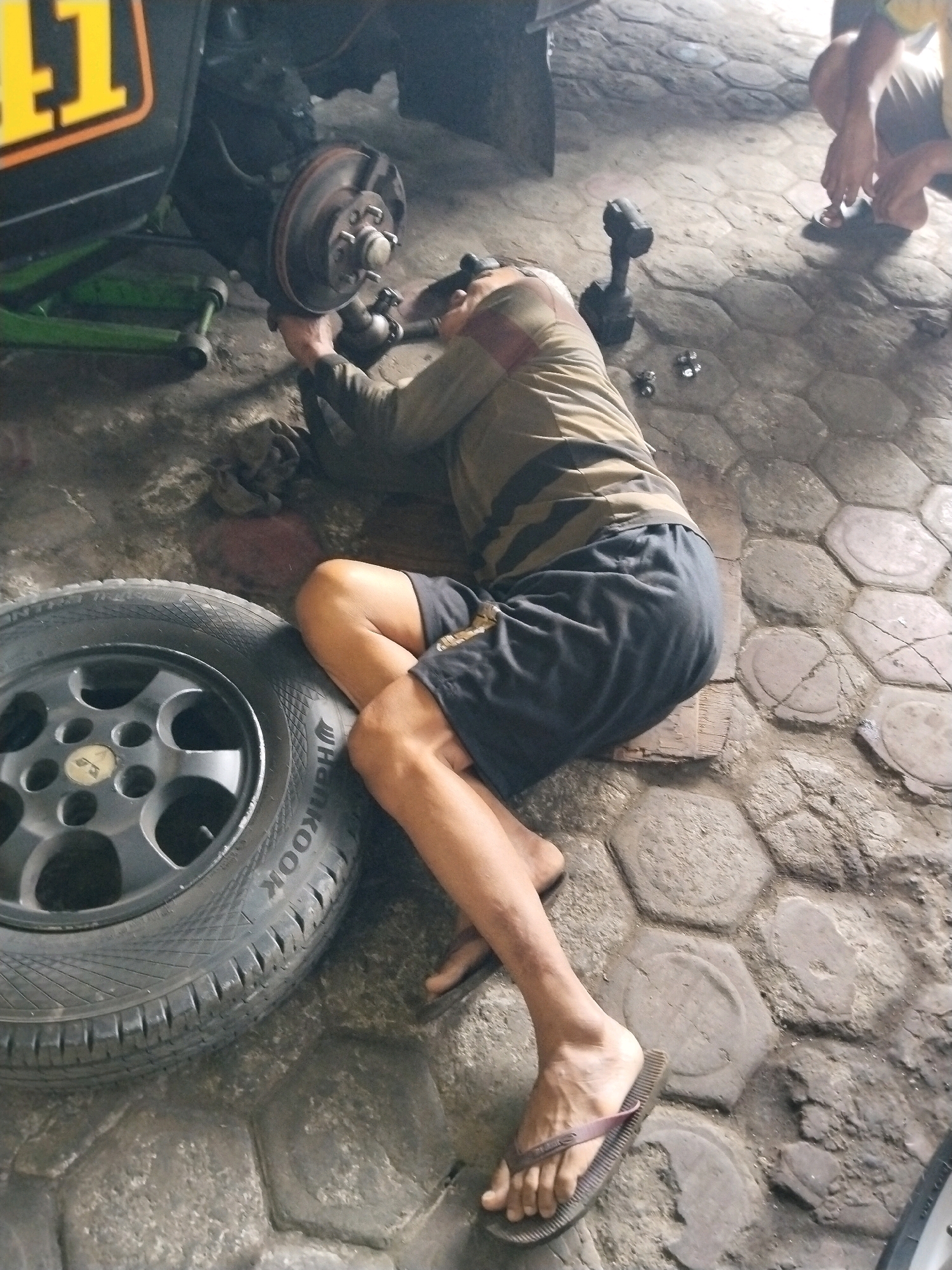In Prigi, Indonesia, a person is working on a vehicle's brake system, lying on their side on a paved ground surface. They are wearing a striped shirt and dark shorts, with a flip-flop on their left foot. A car tire is positioned to the left of the person, and a vehicle's brake rotor and suspension components are visible. A tool, possibly a power drill, lies on the ground nearby, along with other small objects. The environment appears to be an outdoor repair area or a mechanic's workshop.