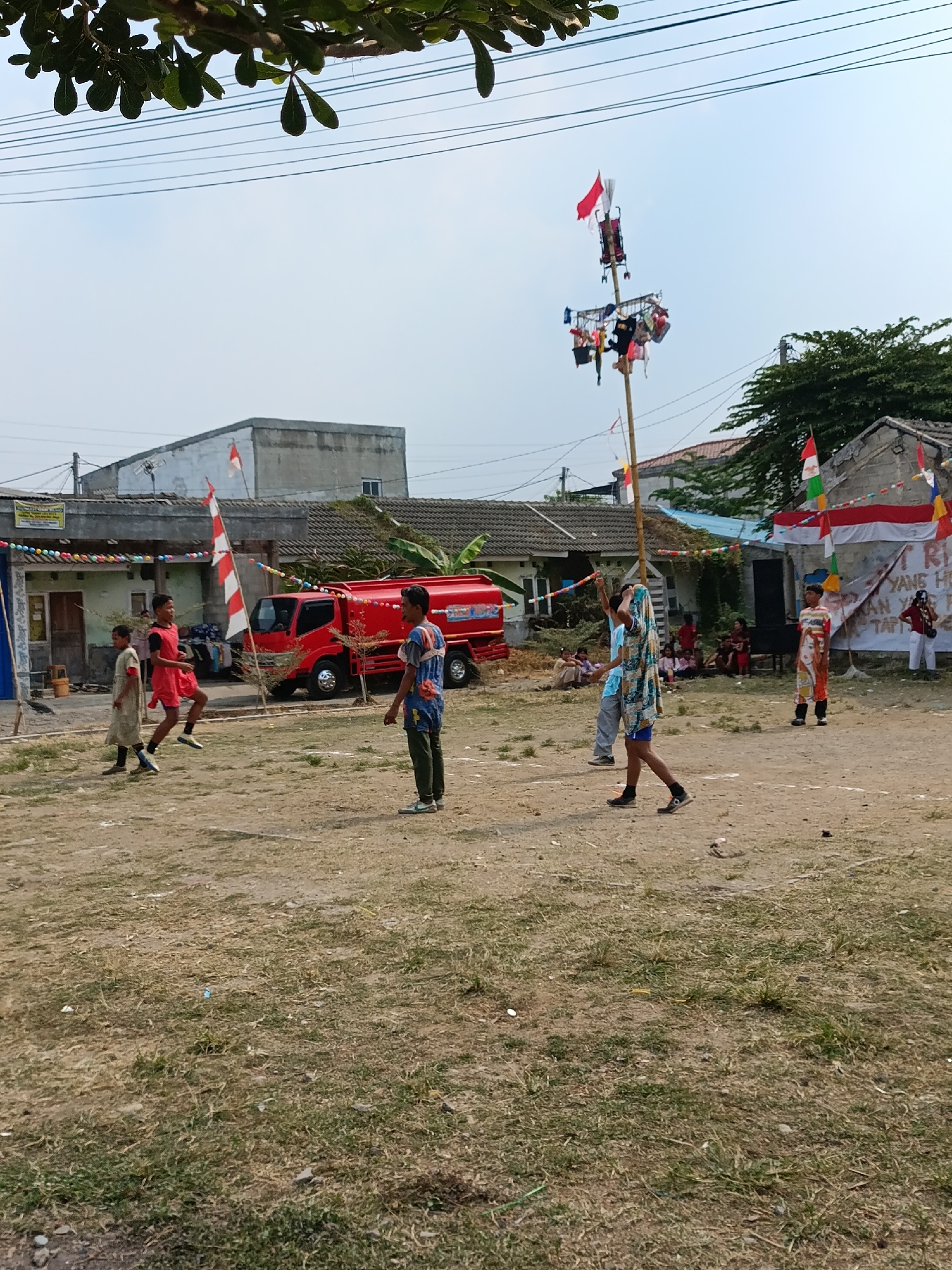 In Cariu, Indonesia, this image captures an outdoor gathering in a dusty, open area, likely a field or a courtyard, with dry grass patches. In the background, several modest, single-story buildings with tiled roofs are visible, some adorned with Indonesian flags and festive banners. A bright red truck, possibly a fire truck or a delivery vehicle, is parked prominently.
The foreground is populated by several individuals, primarily young boys, dressed in casual clothing, some participating in what appears to be a game or a performance. One boy in a red shirt is mid-jump, while others stand or observe. A tall bamboo pole, decorated with flags and what seem to be small offerings or prizes, stands in the center, suggesting a cultural event or competition. The atmosphere appears to be lively and communal, with spectators seated in the background. The sky is clear and bright, indicating daytime.