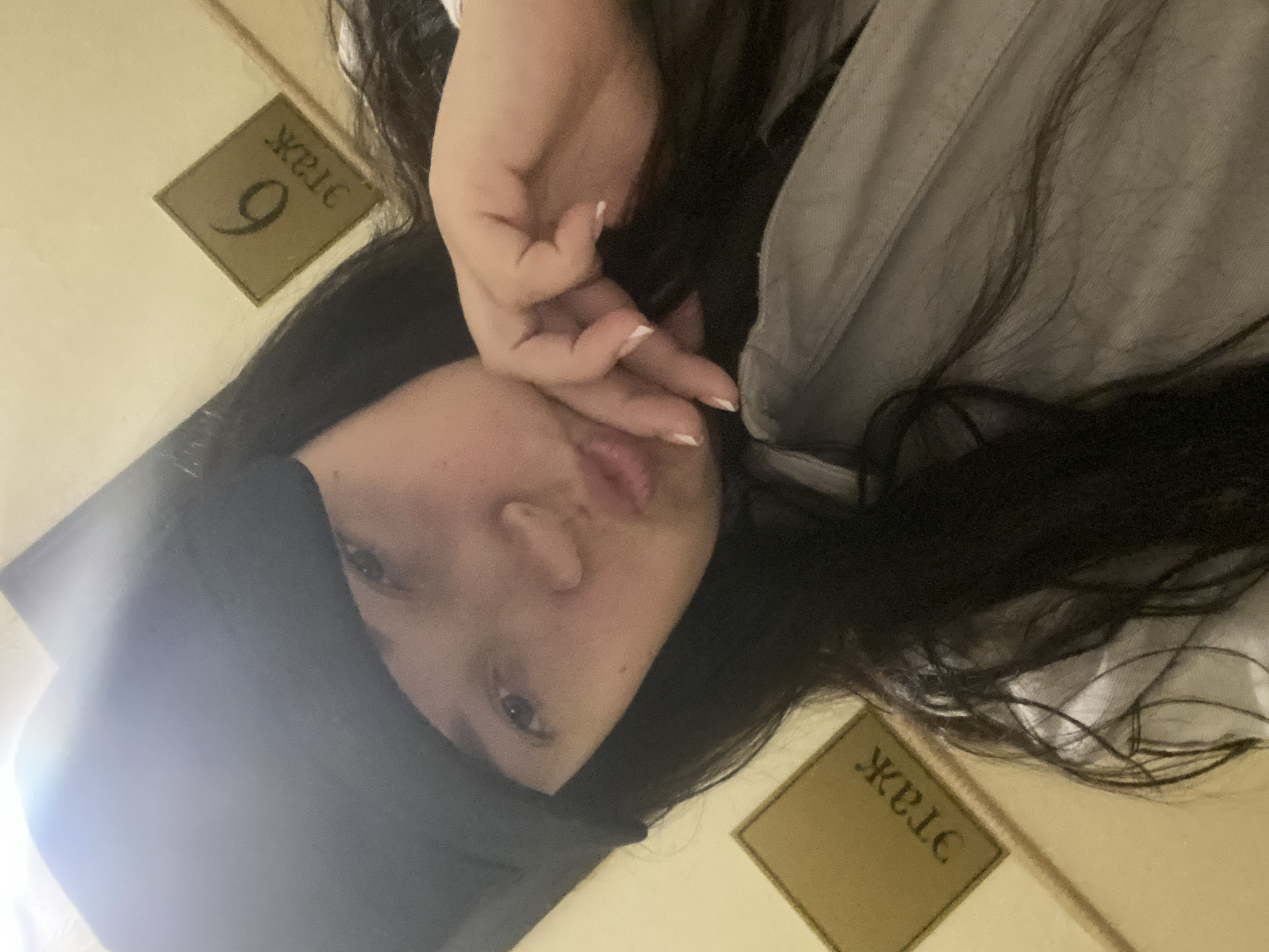 A young woman with long, dark hair is taking a selfie in what appears to be the stairwell of a building in Sochi, Russia. She is wearing a dark hat and a light-colored shirt or jacket. Her hand is raised near her face, partially obscuring it. The number "9" is visible on a sign in the background, along with the Cyrillic word "ЭТАЖ," which translates to "floor." The sign indicates the 9th floor of the building. The lighting is somewhat dim.