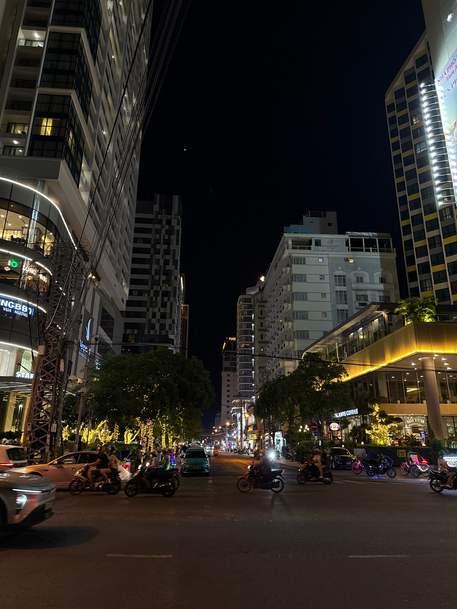 The image captures a nighttime street scene in Nha Trang, Vietnam, likely on a major boulevard. Tall, modern buildings with illuminated windows line both sides of the road. On the left, a building features a curved, illuminated facade, with visible businesses like "VINAGROUP" and "STARBUCKS" on lower levels. A prominent power pole with numerous cables stands in the foreground, obscuring parts of the building on the left. On the right, another tall building displays a large illuminated billboard with text that includes "NHÀ PHỐ." The street is active with vehicular and pedestrian traffic. Several motorbikes, carrying one or two riders, are a dominant feature, navigating the road. Cars and a greenish taxi are also visible. Trees adorned with twinkling lights are planted along the sidewalks, adding to the festive nighttime atmosphere. The sky is dark, indicating it is well past sunset. The overall impression is of a bustling urban environment at night.