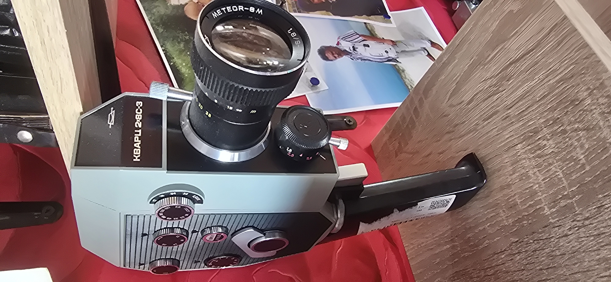 The image shows a close-up, slightly angled view of a vintage, light blue and black film camera with a large, silver and black lens. The camera has a black handle extending to the right. It rests on a red surface, possibly fabric, with several photographs scattered around it. One photograph in the background shows a person in a white and striped outfit, lying down on a blue surface. The camera has several dials and buttons on its body. The lens is labeled "METEOR-8M 1,8/9-36". The body of the camera also has text that appears to be in Cyrillic, reading "КВАРЦ 2-8С-3". The setting appears to be indoors, possibly a home or a shop display, given the presence of the camera and photographs. There are also hints of wooden surfaces in the background, suggesting furniture or shelving. The overall impression is that of an aged piece of equipment, possibly being showcased or used for display. The context provided indicates the location is Gostagayevskaya, Russia. No specific time of day, weather, or discernible emotions are evident.