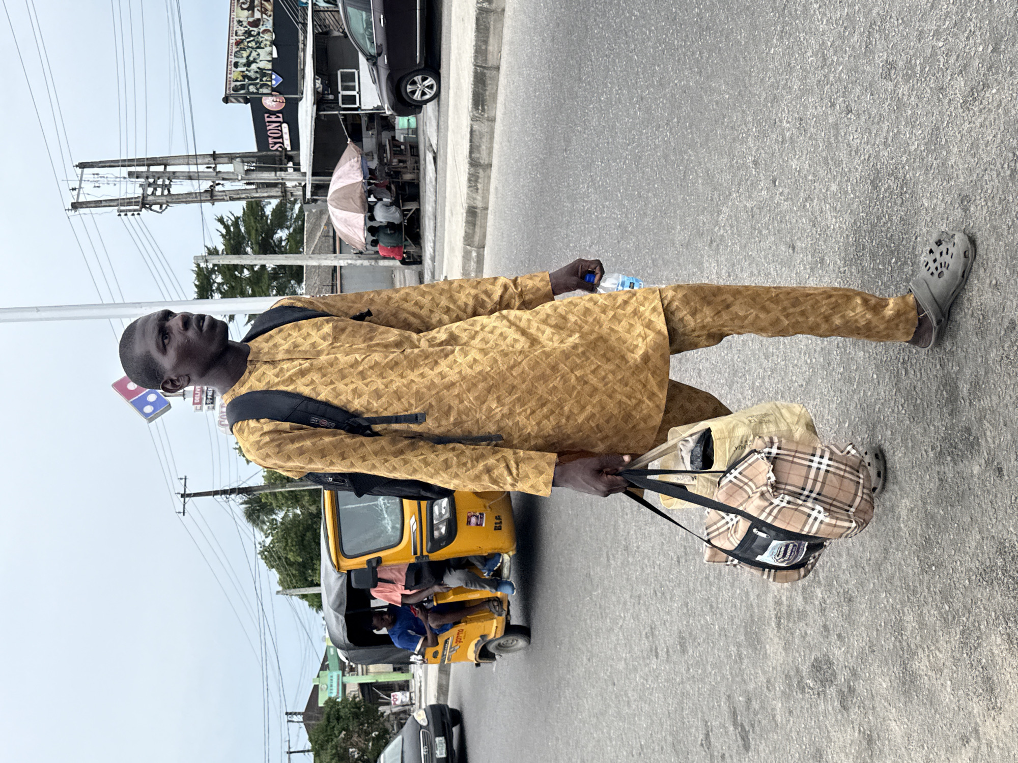 A man walks on the side of a paved road in Ado, Nigeria. He carries a plaid bag in his left hand and a water bottle in his right. He wears a tan, textured, long-sleeved shirt and matching pants, a black backpack, and grey slip-on shoes. A yellow motorized tricycle with passengers passes him on the road. In the background, there is a business with a "STONE" sign, power lines, a Domino's Pizza sign, trees, other buildings, and vehicles.