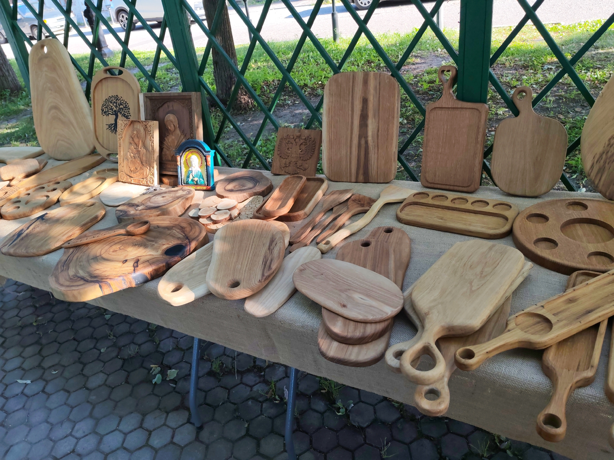 The image displays a table laden with wooden cutting boards and other wooden crafts, likely at an outdoor market or fair. The scene is set under a green lattice structure, with trees and grass visible in the background, suggesting an outdoor, possibly park-like setting. The time of day appears to be daytime, with sunlight casting shadows and highlighting the textures of the wood. The weather seems pleasant, with the sunlight indicating a clear day.
The objects for sale are diverse, featuring various shapes and sizes of cutting boards, some with decorative carvings. Among the items, there are also carved wooden religious icons, including one with an image of the Virgin Mary and another with what appears to be a depiction of a saint. A small, brightly colored icon in a blue and gold frame is also present. Some of the cutting boards have handles or cut-out shapes. There are also smaller wooden items, such as round slices of wood and wooden utensils.
No people are clearly visible in the image, though the presence of a table and a display of goods implies a vendor and potential customers. The overall impression is one of artisanal craftsmanship and a marketplace atmosphere. The text "ПЛИС" is visible on the small, brightly colored icon, which likely refers to the location of Plyos, Russia, as indicated by the context.