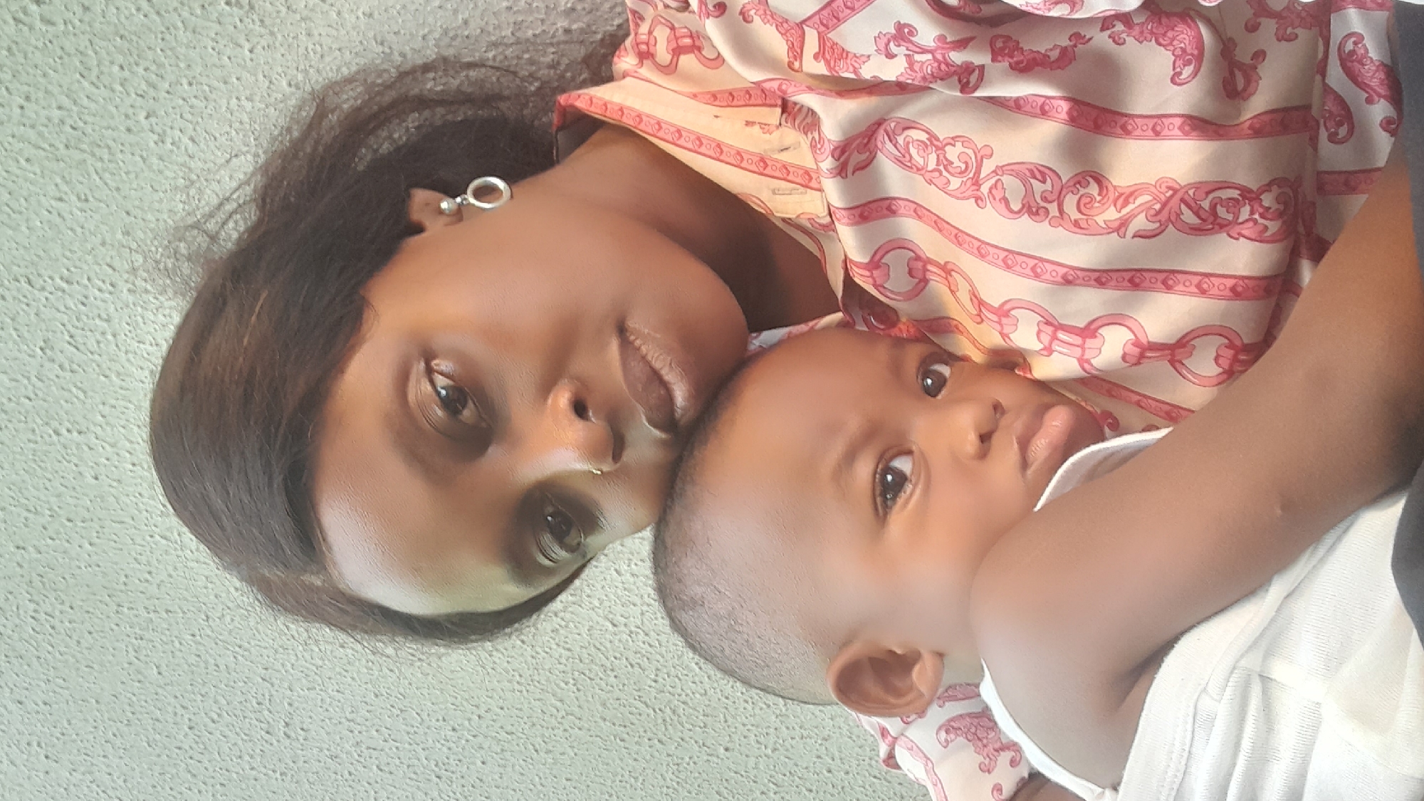 This close-up portrait features a woman and a baby, likely a mother and child, positioned intimately together. The image is oriented vertically, with both subjects looking towards the camera.
The woman, on the left, has dark skin and dark hair that partially covers her forehead. She is wearing a light-colored top patterned with intricate pink or red chain and scroll motifs. A silver-toned hoop earring is visible in her left ear, and she has a small, gold-toned nose ring. Her expression is calm and direct.
The baby, nestled against the woman on the right, also has dark skin and short, dark hair. The baby wears a simple, light-colored or white garment, possibly a onesie or vest. One of the baby's hands is raised, with fingers or a thumb near its mouth. The baby's eyes are wide and focused on the viewer, conveying a serene gaze.
The subjects' heads are close, suggesting a tender bond. The background is a uniform, light-colored, textured surface, possibly a wall or fabric, which keeps the focus entirely on the figures. The lighting is soft and even, indicating a bright, possibly indoor, environment during the day. No text is visible in the image. The photo depicts a moment of quiet connection, originating from Ojomu, Nigeria.