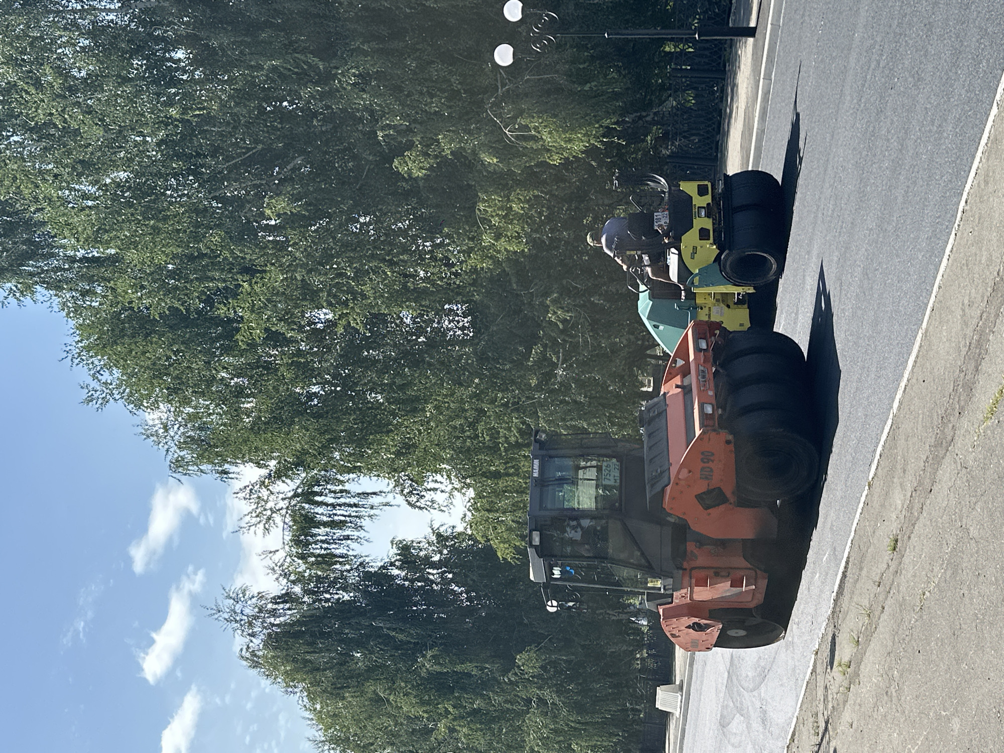 Two road rollers are on an asphalt road, likely undergoing resurfacing. The larger, orange Hamm HD 90 roller is in the foreground, with its cabin visible. Behind it, a smaller, yellow and green Hamm roller is operated by a person. Trees line the street, and the sky is blue with scattered clouds. The scene appears to be a public street in Biysk, Russia, given the context provided.