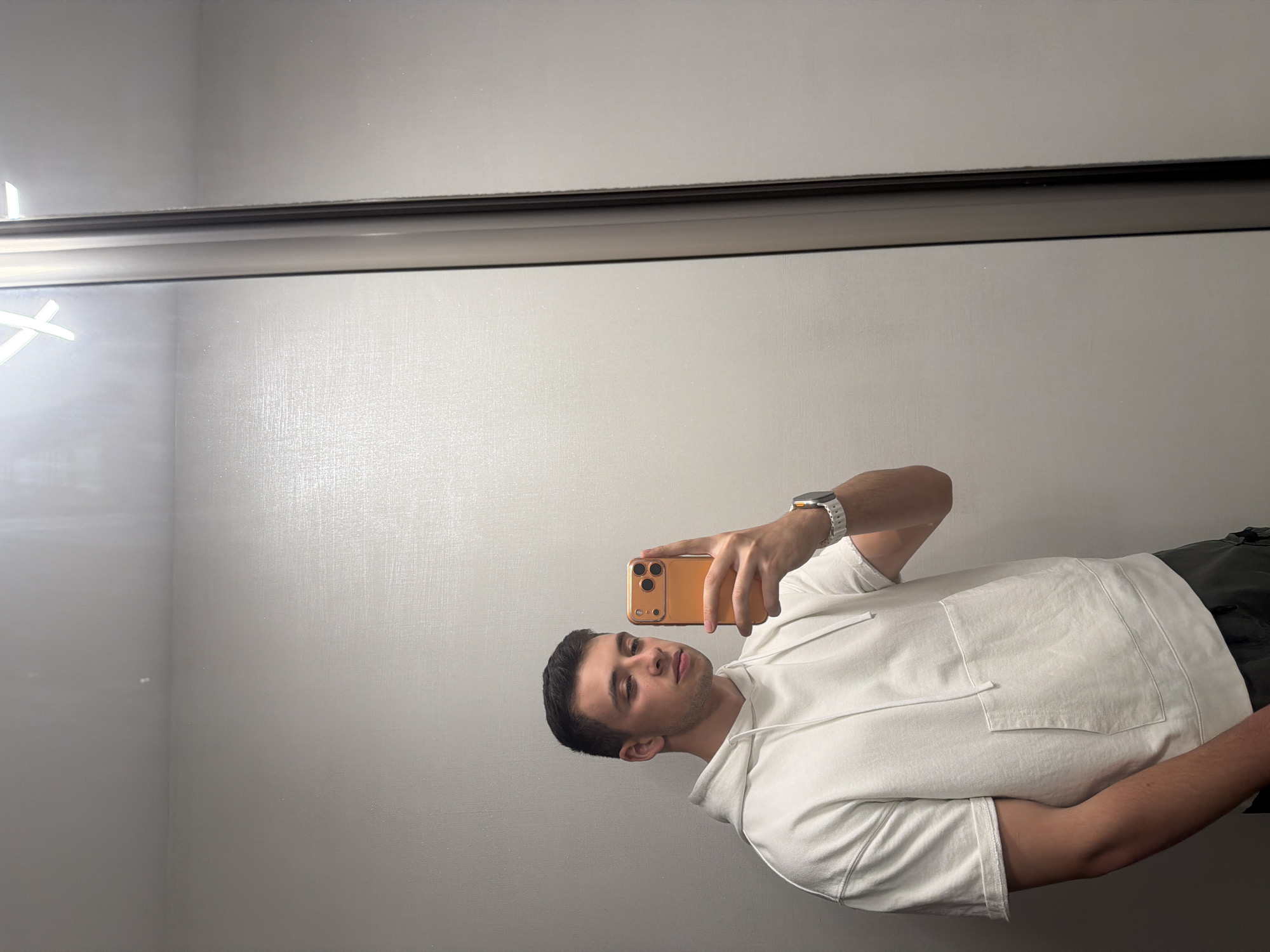 A young man takes a mirror selfie in an indoor setting. He is positioned slightly off-center to the right, looking directly at his smartphone's screen, which is reflected in the mirror. He holds a gold or orange-colored smartphone with three visible camera lenses in his right hand. On his left wrist, he wears a white watch with an orange accent on the dial or crown.
The man is dressed in a white short-sleeve hoodie with a front pocket and drawstrings. He appears to be wearing dark pants or shorts. His expression is neutral, and he has short dark hair.
The background visible in the reflection is a light grey or off-white wall with a subtle, textured finish, resembling a fine weave or stucco. A prominent dark horizontal strip or frame runs across the upper portion of the mirror, likely separating sections of a wall or a door. In the upper left corner of the reflected background, there's a bright, almost star-shaped glare or light reflection. The lighting suggests an artificial indoor environment. There is no visible text in the image.
