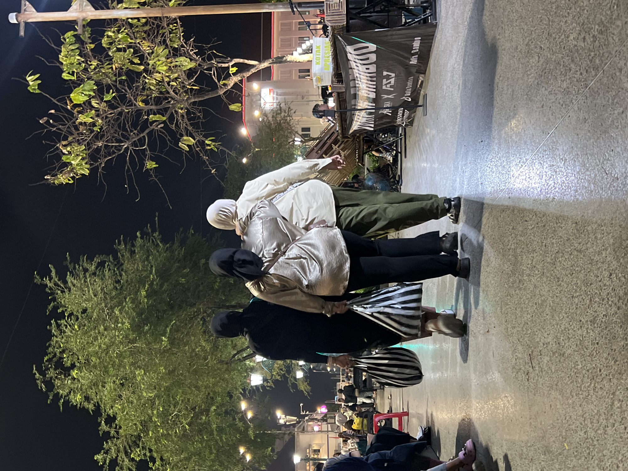 Three individuals, dressed in hijabs, walk away from the camera on a paved walkway at night. The person on the left wears a black dress and carries a black and white striped tote bag. The middle person is in a shimmering gold top and black pants, also holding a black and white striped tote bag. The person on the right wears a cream-colored shirt and olive green pants. Trees with green foliage are visible in the background, illuminated by streetlights. In the distance, buildings with lights on are visible. A banner with "KOTAK APRESIASI" and other text is displayed on the right side of the image, along with a stage setup. The scene appears to be in a public outdoor space in Yogyakarta, Indonesia, possibly a street or plaza area where an event is taking place.