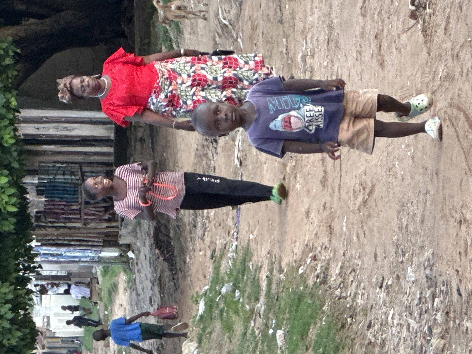 In the city of Narayi, Nigeria, a dirt path winds through a sparse residential area. Green foliage drapes from trees overhead, casting dappled shadows on the ground. A young boy, dressed in a blue t-shirt with a shark graphic and tan shorts, walks towards the viewer, his expression neutral. To his right, a woman in a vibrant red top and a floral skirt stands looking forward, her hair styled in an elaborate updo. To the woman's left, a girl in a striped t-shirt and black pants walks away from the viewer, her gaze turned to the side. Another boy, wearing a blue t-shirt and dark shorts, walks ahead of the girl, carrying a red bag in his left hand. In the background, several buildings with corrugated metal roofs and wooden structures line the street. A tan dog, standing in the distance, appears to be looking away from the camera. The overall scene is one of everyday life in a Nigerian community.