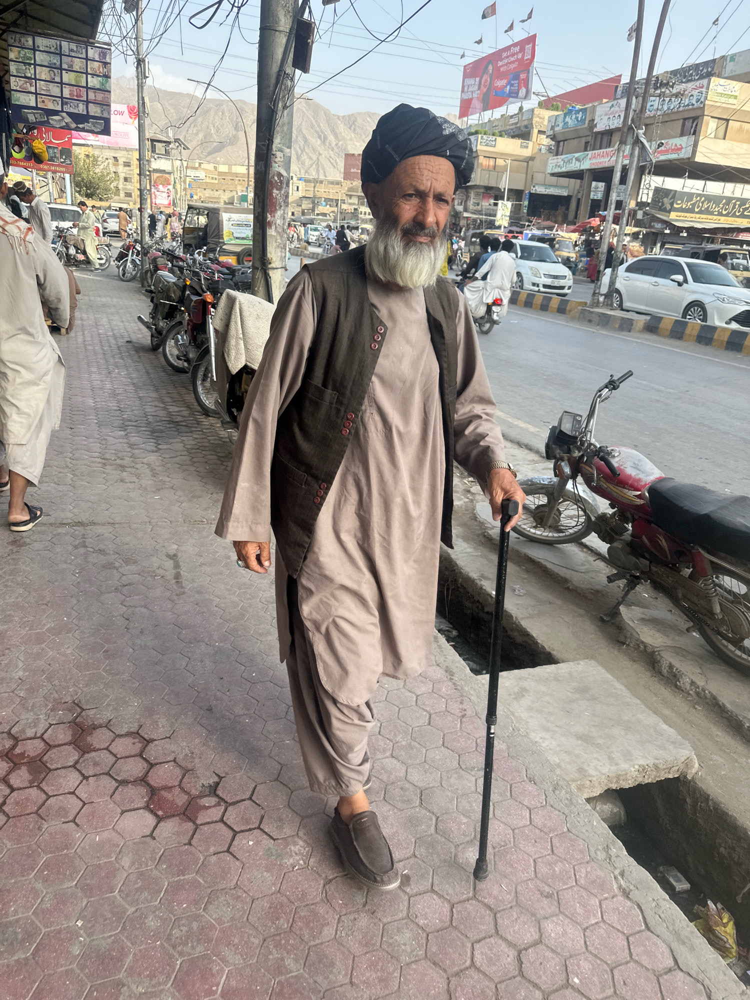 An elderly man with a long white beard and a black turban walks with a cane on a sidewalk in Quetta City Tehsil, Pakistan. He wears a brown tunic and a dark vest with buttons. To his left, other pedestrians and motorcycles are visible, and a street with cars runs parallel to the sidewalk. Shops line the street in the background, and mountains are visible in the distance. The sidewalk is paved with hexagonal red tiles, and a ditch runs along its edge. The scene appears to be a busy street market.