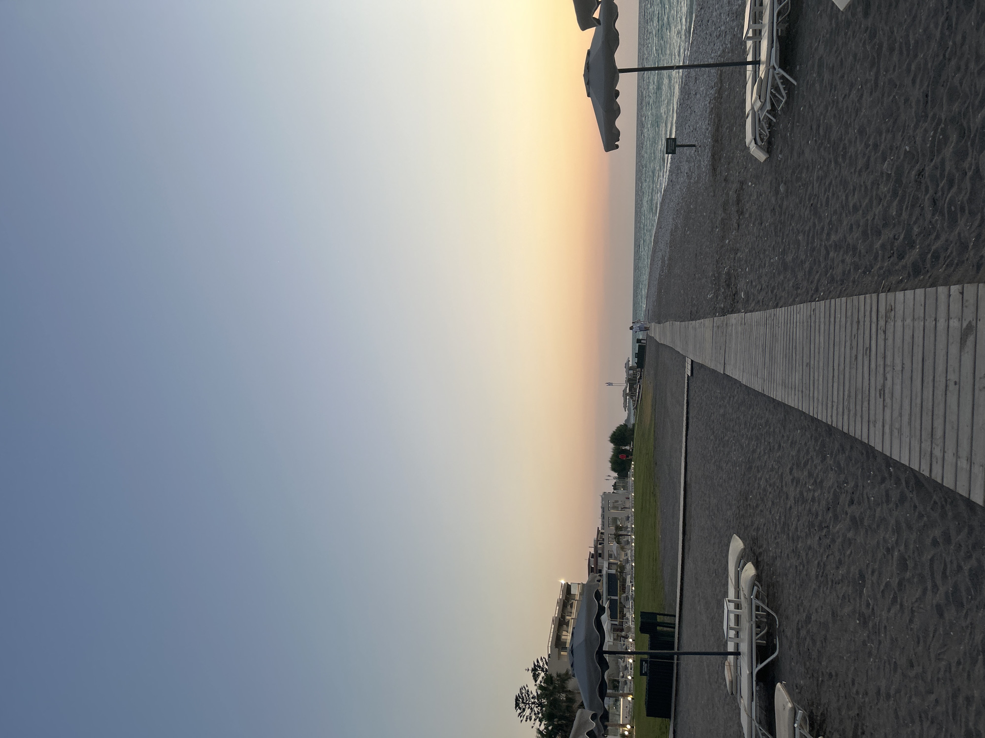 This media file depicts a beach scene at sunset in Rhodes, Greece. The image is oriented vertically, with the sky occupying the left half and the beach and resort on the right. The sky displays a gradient of colors, transitioning from a pale blue at the top to soft orange and yellow hues near the horizon, indicating it is dusk.
The beach itself is covered in dark sand and pebbles. A wooden boardwalk runs along the edge of the beach, providing a path. Several empty white lounge chairs with umbrellas are scattered across the sand, suggesting a resort area. In the background, there are modern hotel buildings with white facades and balconies, surrounded by green lawn areas. Palm trees and other Mediterranean vegetation are visible, adding to the tropical resort feel.
The sea is calm and appears in shades of blue-grey. There is a small, distant structure or building visible on the horizon near the sea. The overall atmosphere is serene and peaceful, characteristic of a quiet evening by the sea. No people are prominently visible in the frame, though the presence of lounge chairs and resort buildings implies human activity and vacationing. No visible text is present in the image.