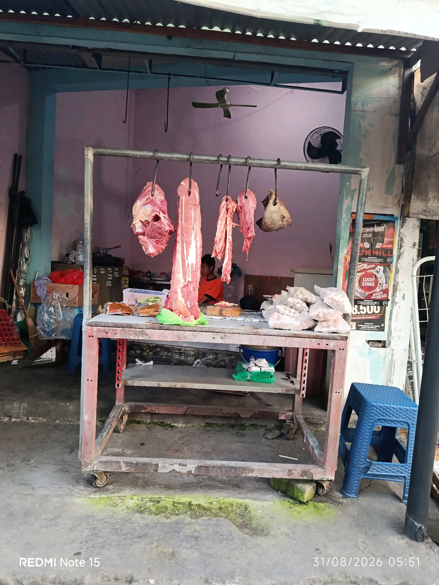 The image shows a meat stall in Keboansikep, Indonesia. In the foreground, a metal cart with two shelves holds various items. On the top shelf, there are several plastic bags filled with what appear to be meatballs, and some packaged food items. A green plastic sheet and other miscellaneous items are also visible on this shelf. The bottom shelf is mostly empty, with a few scattered items and a piece of green material.
Hanging from hooks above the cart are several large pieces of raw meat, including what looks like a whole side of beef. To the left of the cart, there is a broom and some plastic bags with items inside. A blue plastic stool is positioned to the right of the cart.
In the background, the stall is open to a street. A person wearing an orange shirt is visible behind the counter, appearing to be engaged in some activity related to the meat. A ceiling fan is mounted on the wall behind them. The walls of the stall are painted a light pink color. There are posters on the wall to the right of the stall. The ground is concrete and appears somewhat weathered, with patches of green growth. The overall scene depicts a typical local market setting.