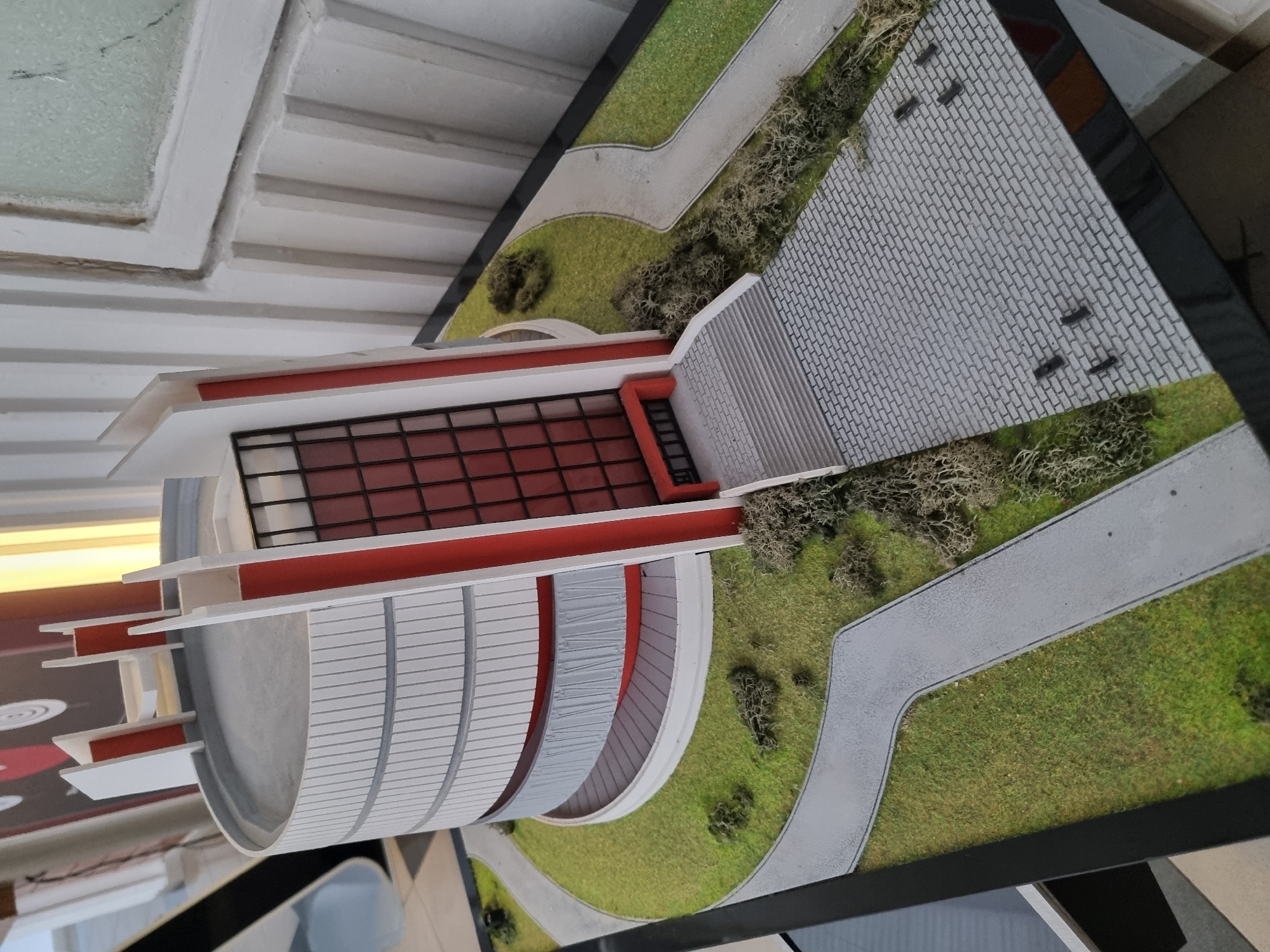 A high-angle shot shows an architectural model of the Panorama Pleven Epopee 1877 museum located in Pleven, Bulgaria. The model sits on a black, rectangular platform and includes miniature representations of the museum building, surrounding landscaping, and walkways. The museum is depicted with its distinctive semicircular white facade, trimmed with red accents. The windows are filled with a dark red hue. The landscaping features green turf and miniature trees. A gray, brick-patterned walkway approaches the museum entrance. A gray, winding path is visible near the bottom-right corner of the model, surrounded by green areas. The background includes a white ceiling and a partial view of an interior with an overhead light and possibly other objects.