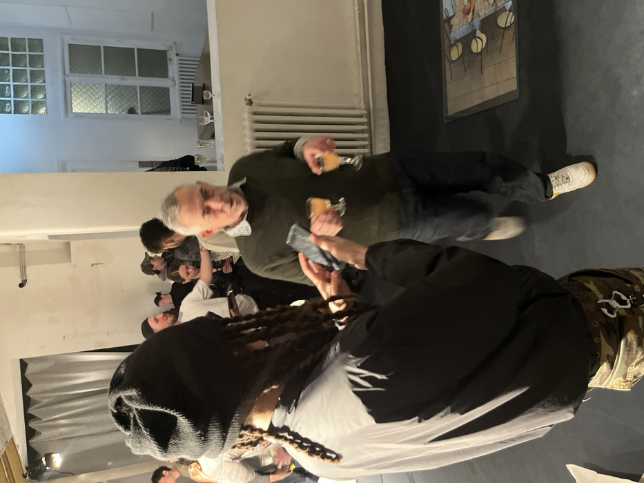 An indoor scene depicts a social gathering or event in an urban setting within Paris, France. In the foreground, a person with dreadlocks, wearing a black beanie, a black long-sleeved top, and camouflage pants with two white carabiner clips attached, is seen from behind. They are holding a dark smartphone horizontally with both hands, pointed towards the mid-ground.
To the right, a middle-aged man with short grey hair, wearing a dark green crew-neck sweater over a patterned collared shirt, dark blue jeans, and light-colored athletic shoes with yellow soles, is captured in motion, slightly blurred. He has his mouth partially open and is holding two stemmed glasses, each containing a light amber liquid.
The background shows additional individuals mingling in various states of focus, some holding drinks. The environment features white walls, a high ceiling, and dark flooring. Architectural elements include a tall white column radiator on the right wall, a recessed window with multiple square glass blocks in its upper section, and a frosted or patterned lower glass section. A light-colored wooden table with several empty glasses is visible beneath this window. On the floor in the mid-right, a rectangular embedded display or artwork with circular motifs is visible. A light-colored curtain is partially visible in the far left background. The space is brightly illuminated, primarily by artificial light.