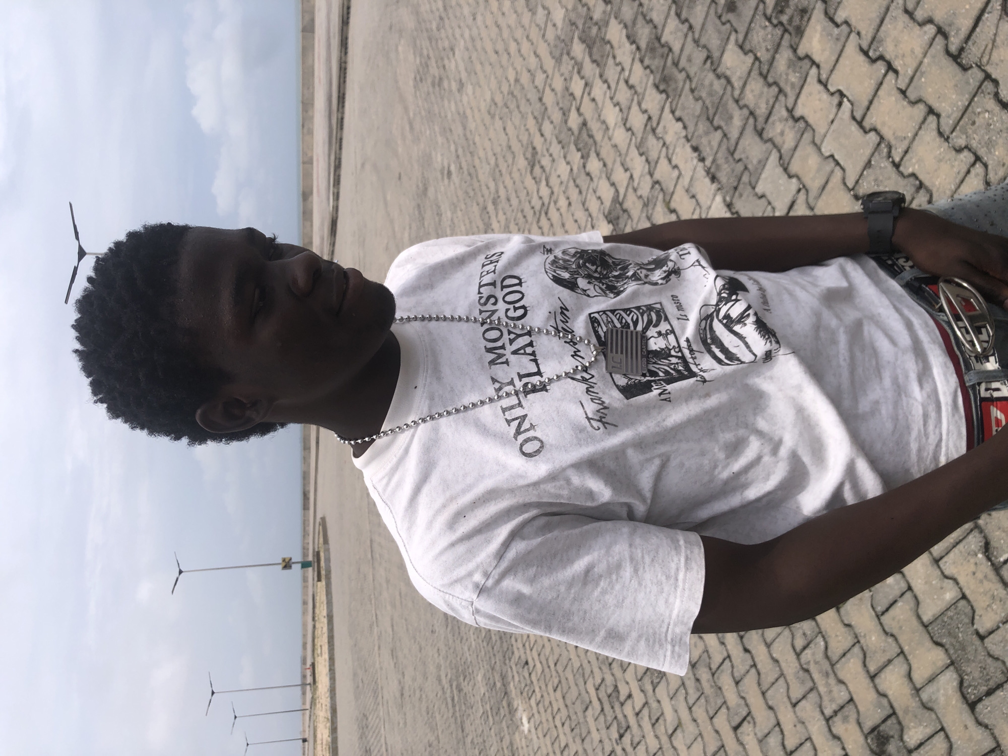 A young Black male stands outdoors on a paved surface. He is wearing a white t-shirt with a graphic print, a silver chain with a pendant, a watch on his left wrist, and jeans with a belt. The background shows a cloudy sky and streetlights along a body of water. The location is identified as Ikoyi, Nigeria.