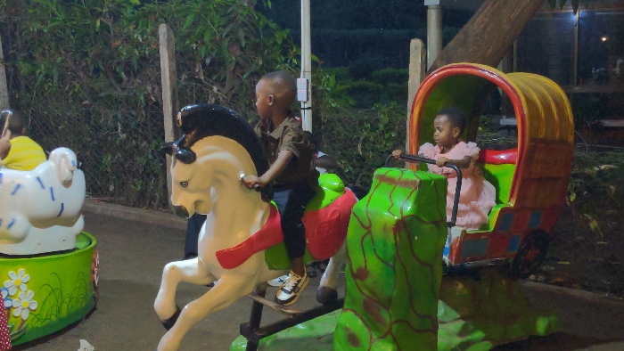 The image displays an outdoor children's amusement area during evening hours in Moshi, Tanzania. Two young children are visible on stationary rides. On the left-center, a male child with short dark hair, wearing a brown collared shirt, dark trousers, and white-and-black sneakers, is seated on a white toy horse with a black mane and red saddle, mounted on a green base. He grasps the horse's handles. To the right, a female child with dark hair, dressed in a pink frilly dress, occupies a multi-colored carriage-style ride featuring a yellow roof, a green and brown textured base, and checkered side patterns. She holds an internal bar. A third person, wearing a yellow shirt, is partially visible on the far left, positioned near a white lamb-shaped ride with blue markings and a green base. The ground surface beneath the rides is paved. The background consists of dense dark green foliage, several vertical wooden posts, and sections of a chain-link fence, all illuminated by artificial light sources. An electrical utility box is affixed to a wooden post behind the male child's ride, and a large tree trunk is present in the upper right background.