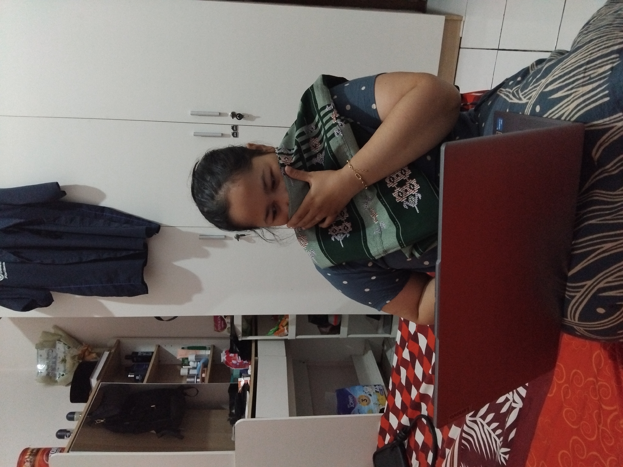 In a domestic setting, likely in Sambirata, Indonesia, a woman is seated on a bed covered with a red and white checkered bedspread. She is using a red laptop, which rests on her lap. The woman is wearing a blue polka-dot shirt and a green scarf adorned with intricate white and black patterns, which she is using to cover the lower half of her face. A gold bracelet is visible on her right wrist. The background shows a white wardrobe with two doors, one of which has a key lock. To the left of the wardrobe, a shelf unit is visible, holding various items including toiletries, a black hat, and a backpack. A dark blue garment, possibly a uniform, hangs on the wall above the shelves. The flooring appears to be tiled.