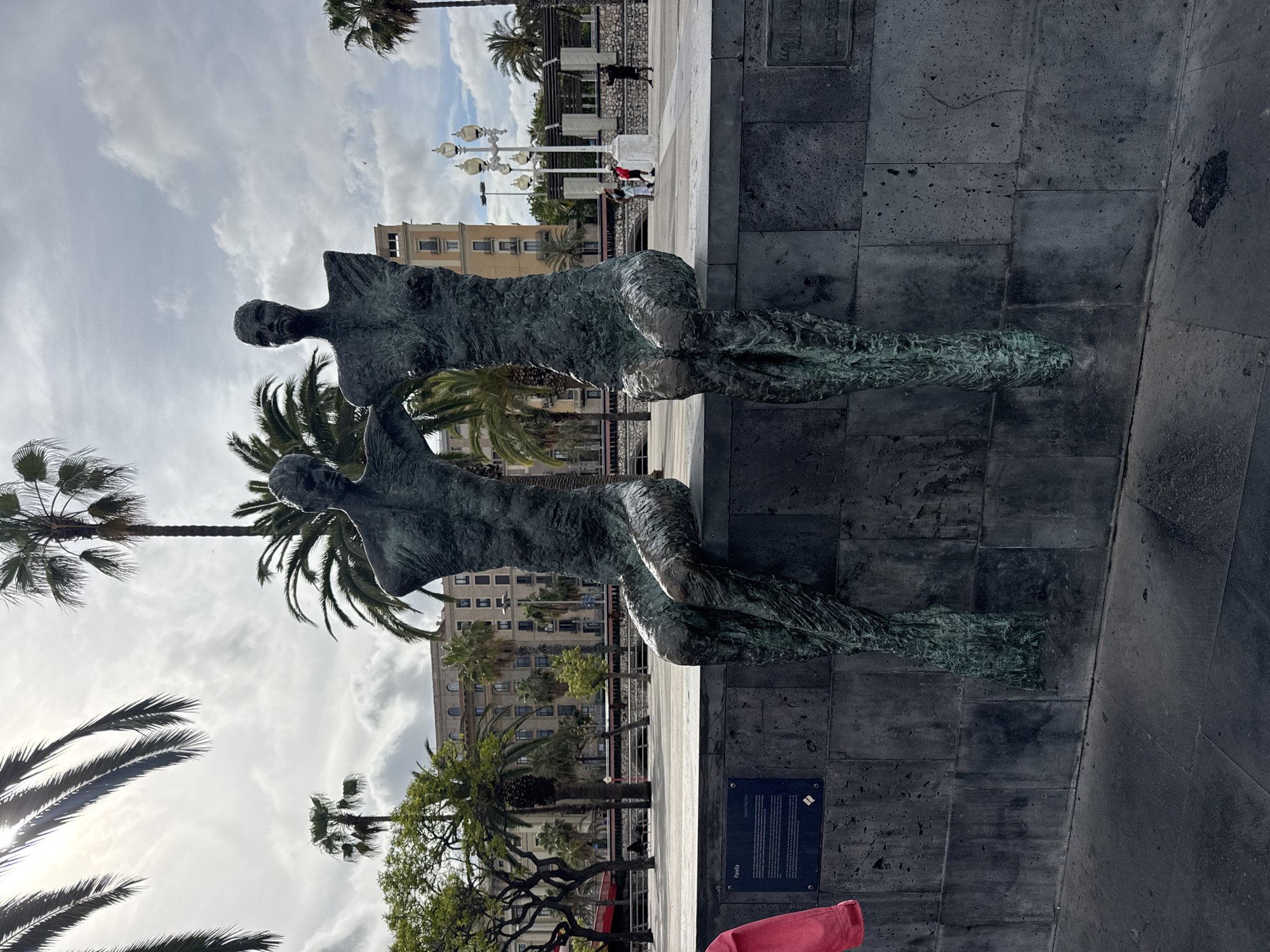 The image depicts a bronze sculpture of two human figures, possibly a man and a woman, on a stone platform. The figures are stylized and abstract. The background features a cityscape with palm trees, buildings, and lampposts. There are several people walking in the background, some appearing to be tourists. The location is likely a public space or park in Barcelona, Spain, based on the architectural style and the presence of palm trees. A plaque with writing is visible on the base of the sculpture. The lighting indicates it's daytime, with a partly cloudy sky. The camera angle is slightly rotated.