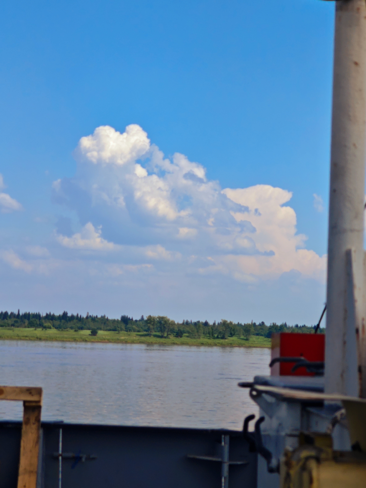 The image depicts a view from a vessel on a body of water, likely a river, with the distant shoreline visible on the horizon. The sky is bright blue with large, fluffy white clouds. The water appears calm, reflecting the sky. The foreground shows parts of the vessel, including a metal railing and some equipment, with a wooden structure on the left. The shoreline is lined with a dense forest of green trees. The overall scene suggests a sunny day in a rural or natural setting. Given the context provided, this scene is likely located in or near Усть-Кемь, Russia, a town situated on the Kema River.