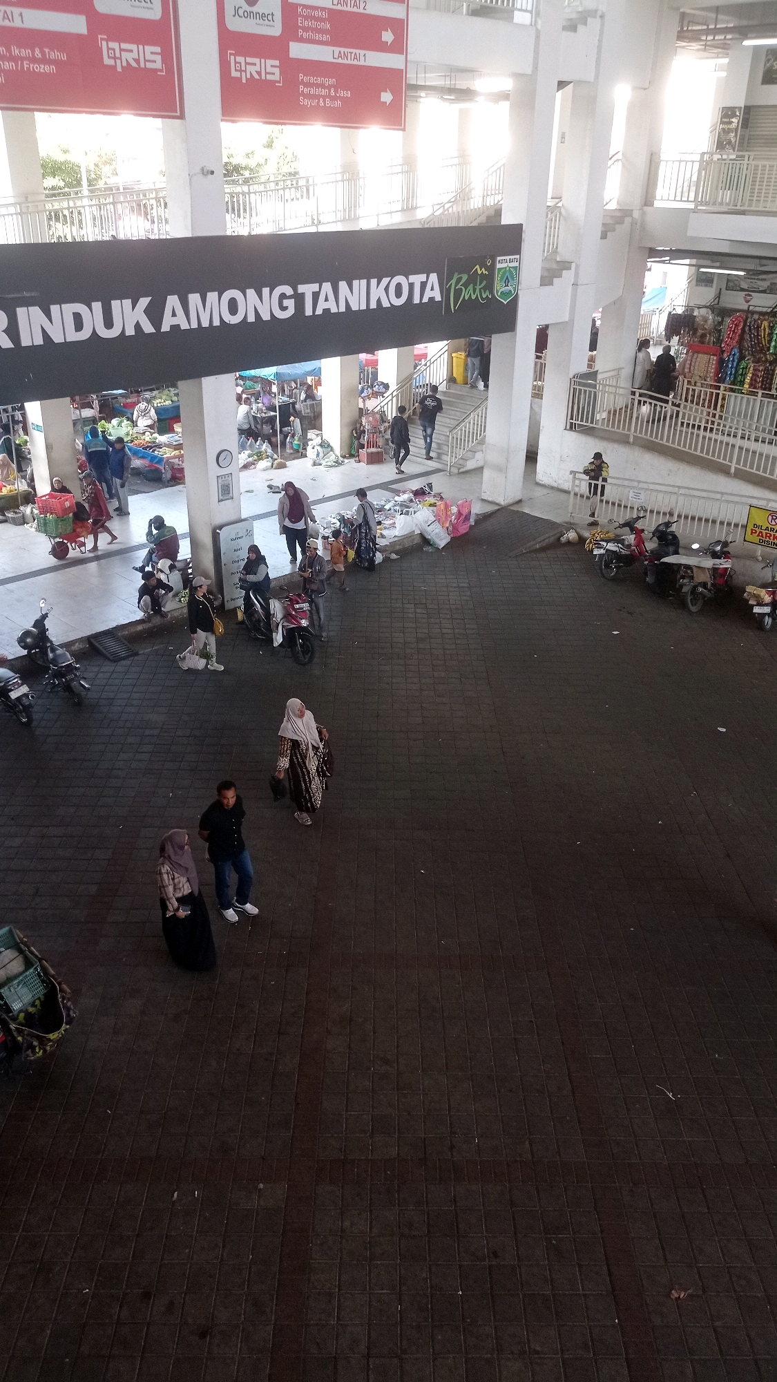 The image depicts a busy market scene in Batu, Indonesia, specifically the "Pasar Induk Among Tani." In the foreground, the paved ground is predominantly dark gray tiles. Several individuals are scattered across this area. To the left, a person in a black hijab and a patterned brown and white robe walks away from the camera. Further back, a person in a black shirt and blue jeans stands facing the camera, while a person in a pink hijab and a dark skirt is to their left.
The middle ground reveals more activity. Several motorbikes are parked, including a red motorbike with a rider. Stalls with various goods are set up, some covered with tarps. People are browsing or interacting near these stalls. In the upper left of the image, a banner reads "PASAR INDUK AMONG TANI KOTA BATU," indicating the location and name of the market. Above this banner, signs display "Lantai 1" and "Lantai 2," suggesting multiple levels to the market.
The background shows the structure of the market, with concrete pillars and elevated walkways. Sunlight streams in from the right, creating bright areas and shadows. More people are visible on the upper levels and near the stairs. The overall impression is of a bustling urban marketplace with ongoing commerce and pedestrian traffic.