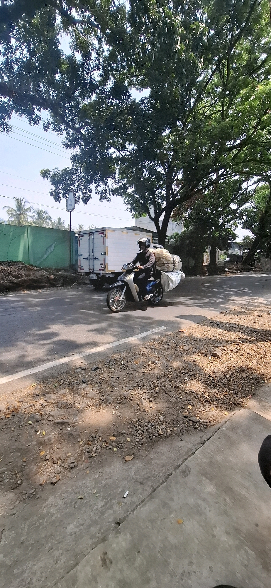 A person is riding a motorcycle on a road in Cisalak, Indonesia. The motorcycle is carrying a large amount of cargo, appearing to be sacks of goods, secured to the rear. The rider is wearing a helmet and dark clothing. A white truck is parked on the side of the road behind the motorcycle. The environment includes a road with a white line, a dirt shoulder with rocks and debris, and a lush green tree canopy overhead. A green wall and some palm trees are visible in the background. The sky is clear and blue.