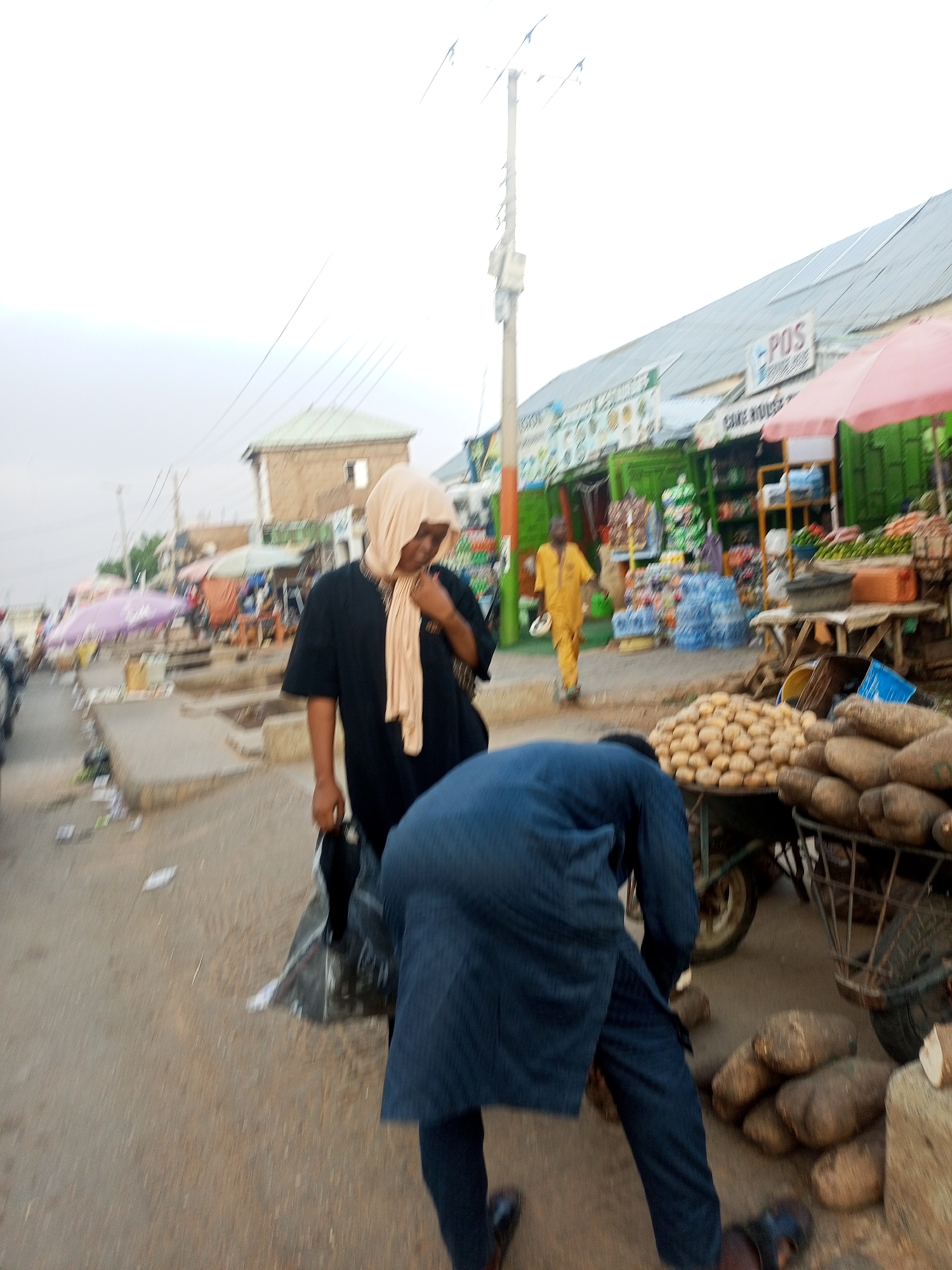 The image depicts a bustling marketplace in Gombe, Nigeria. A woman wearing a hijab and black clothing stands near a man in blue who is picking up yams. Various produce, including potatoes and yams, are displayed on carts. In the background, there's a row of shops with signage, including one marked "POS," and people can be seen walking by. The setting shows a typical urban street with overhead power lines and a mix of buildings.