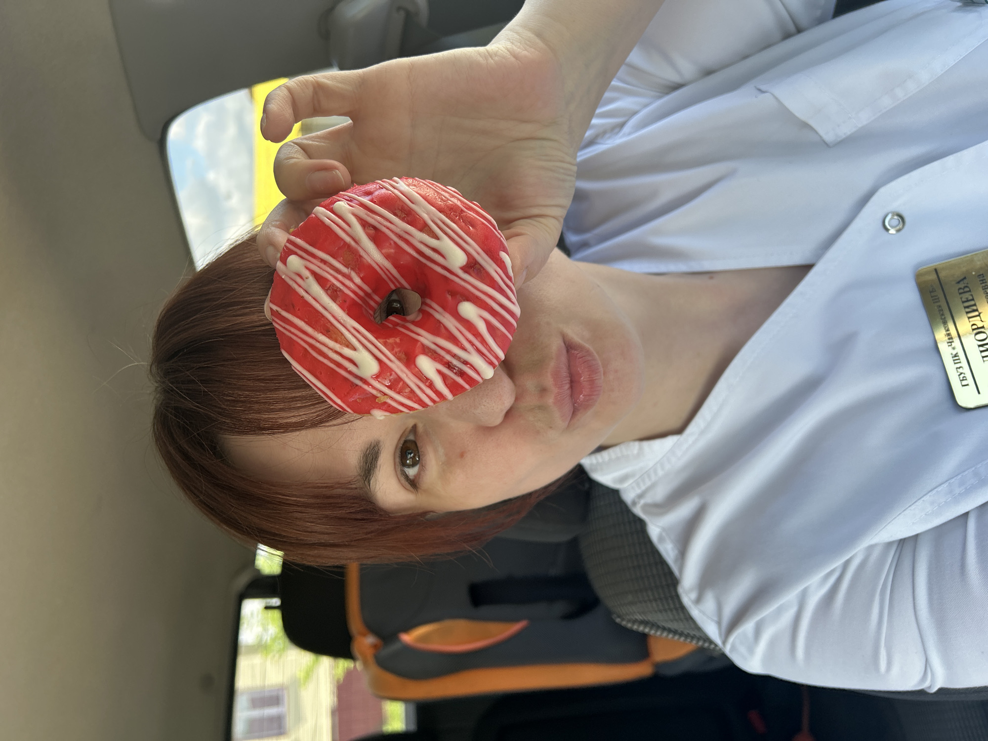 A woman with reddish-brown hair is playfully posing inside a car, holding a bright red donut with white icing stripes up to her face. Her right eye peers through the donut's hole, and her lips are slightly parted in what appears to be a lighthearted expression. She is wearing a white collared shirt, possibly a uniform, and a golden name tag or ID badge is clipped to it.
The visible text on the badge is in Cyrillic, with "МОРГУНОВА" (Morgunova) clearly legible, along with partially visible letters like "ФГБУЗ" and "НБХЗПК". The background shows the car's headliner and a blurred view through the window, revealing a bright sky and some distant outdoor elements, suggesting it is daytime. A car seat, possibly a child's, is visible in the lower left background, further confirming the interior of a vehicle. The scene conveys a moment of lightheartedness.
