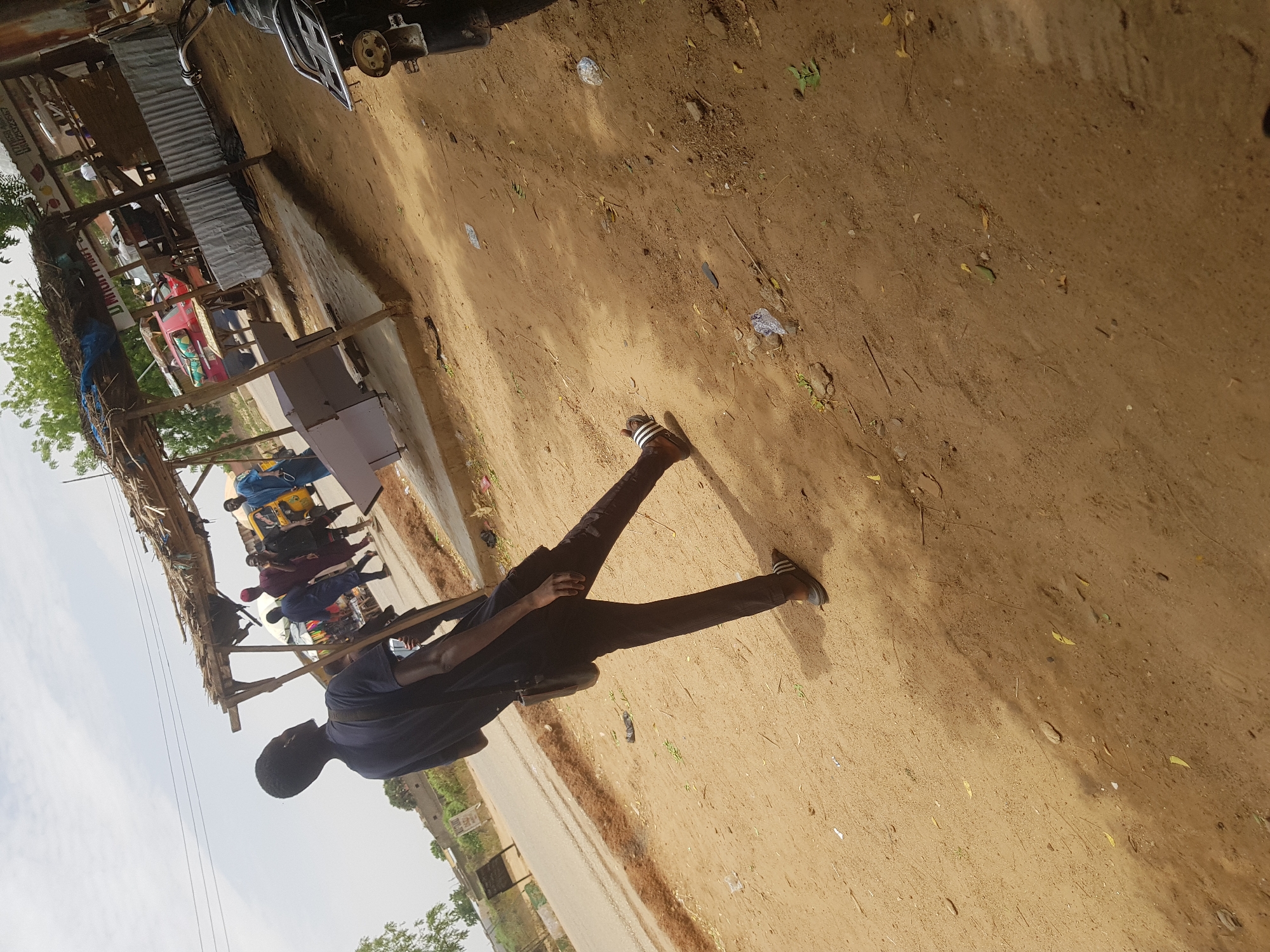 A medium shot captures a man in Dabawa, Nigeria, from a low angle while he is walking on a sandy, dirt path. The man is wearing dark clothing and sandals and appears to be carrying a bag or satchel. He is walking away from the camera along the path. The path extends into the background, where other people are visible near a series of roadside stalls with corrugated metal roofs, a common sight in Dabawa. The environment consists of dirt, foliage, and makeshift structures along the road. The presence of people near the stalls suggests a market or commercial area.