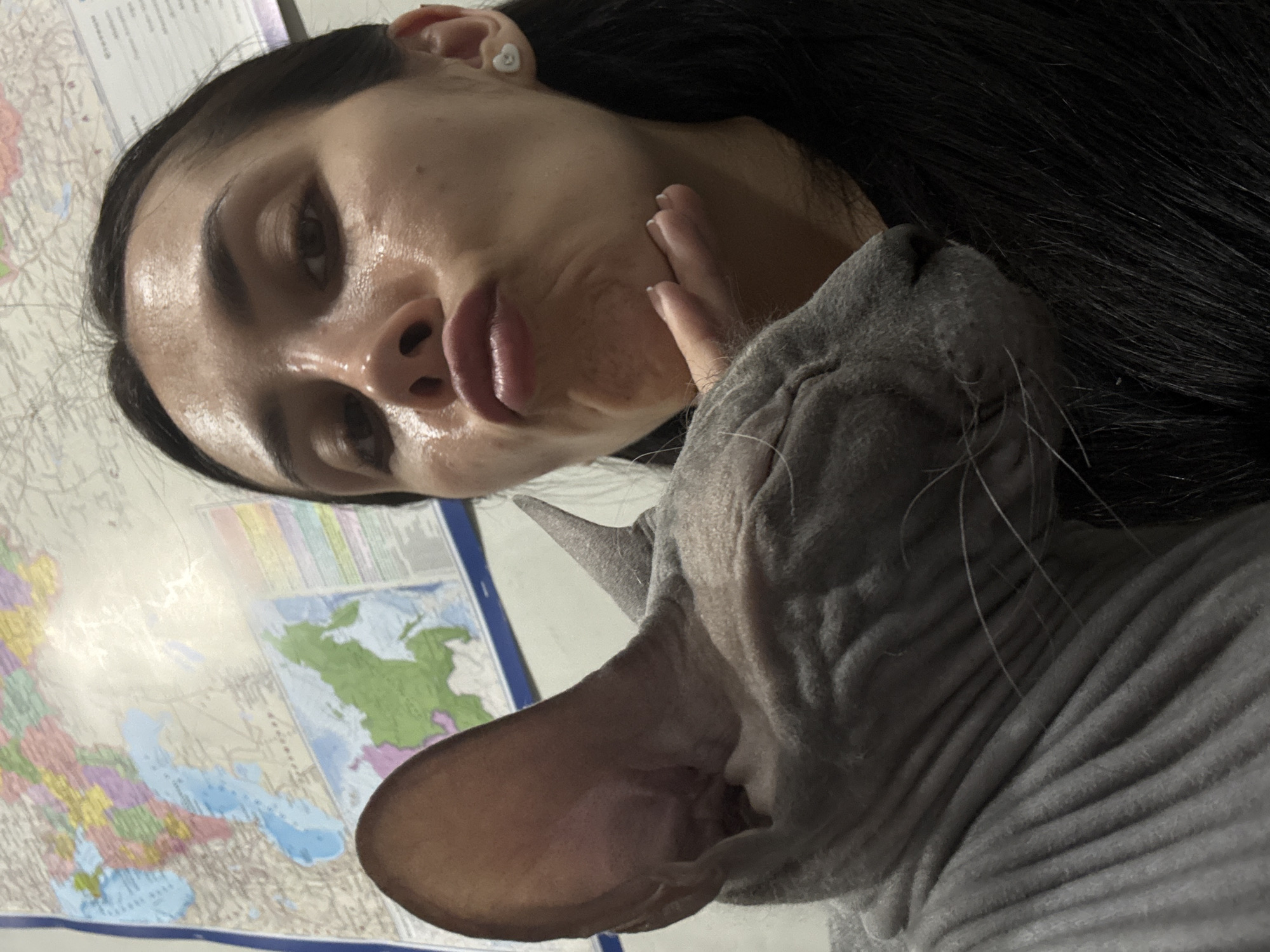 A selfie depicts a woman with long dark hair and a Sphynx cat. The woman, positioned on the left, has moist skin, dark eyes, and pursed lips. Her left hand is raised near her face. The Sphynx cat, on the right, has wrinkled skin and closed eyes. A world map is visible in the background. There is no evidence to suggest a location in the city of Votkinsk, Russia.
