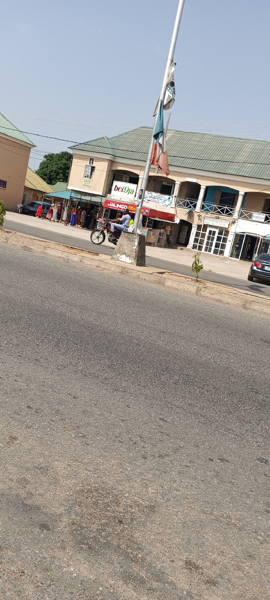 The image depicts a street scene in Jalingo, Nigeria. A man is riding a motorcycle on the right side of the street, moving towards the viewer. Behind the motorcycle, several shops are visible with colorful awnings and signs, including one that reads "bet9ja" and another with "JALINGO WINS". To the left of the shops, a row of women are standing, some wearing colorful dresses, likely vendors or shoppers. Cars are parked on the side of the road near the shops. The street is paved with asphalt, and a concrete curb separates it from the sidewalk. A metal pole with several flags is prominently placed near the center of the image. The sky is clear and blue, indicating a sunny day. The overall impression is that of a busy commercial area in a Nigerian city.