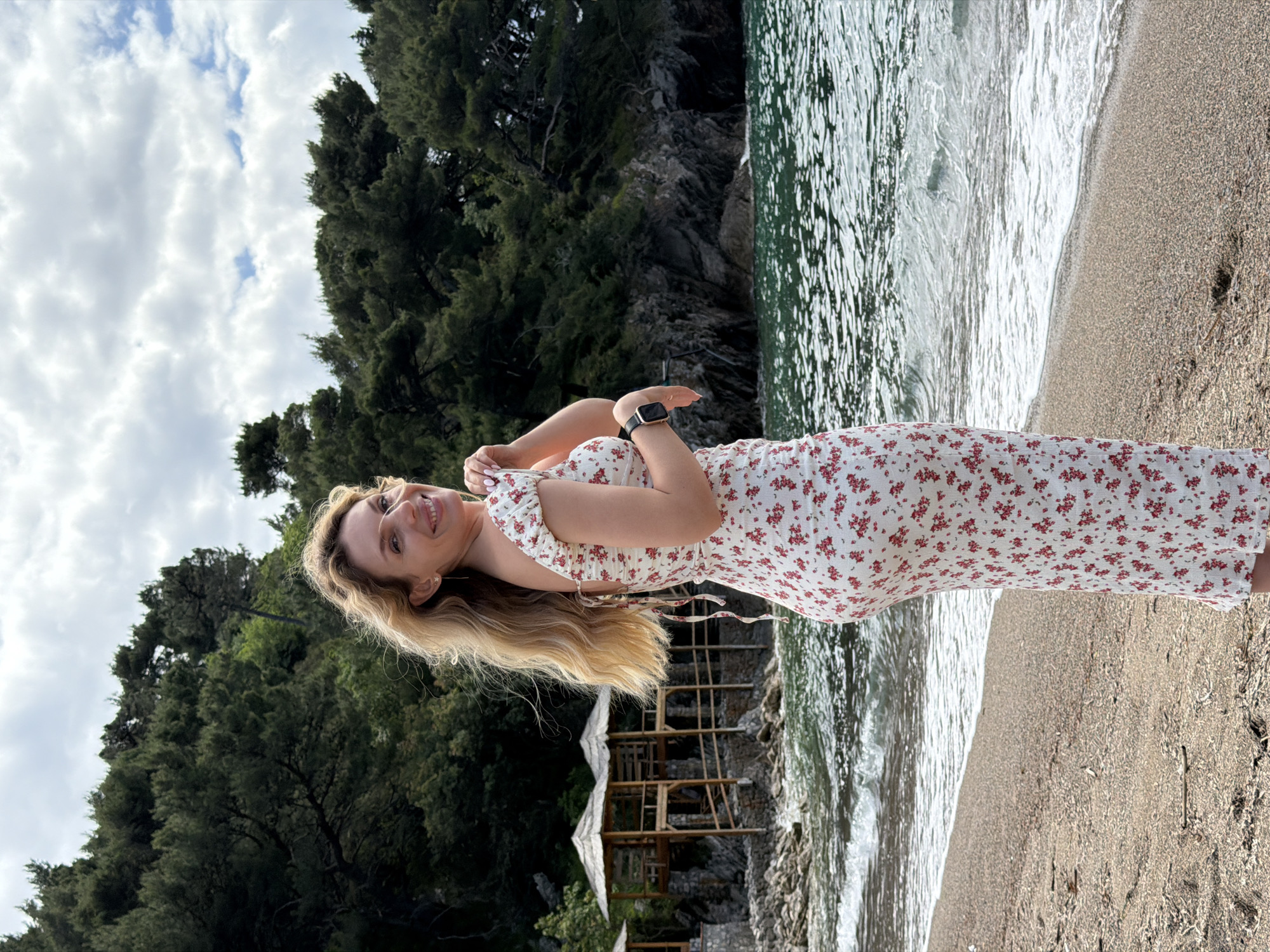 A woman with long blonde hair, wearing a white floral dress and a black smartwatch, stands on a sandy beach. The beach appears to be in Petrovac centar, Montenegro, as suggested by the typical Montenegrin coastline including the rocky hills and lush green trees in the background. She is smiling and looking towards the camera. The beach meets the turquoise sea, with waves gently rolling onto the shore. Behind her, one can see wooden structures, which could be parts of beachside facilities like cabanas or sunshades. The sky is overcast.