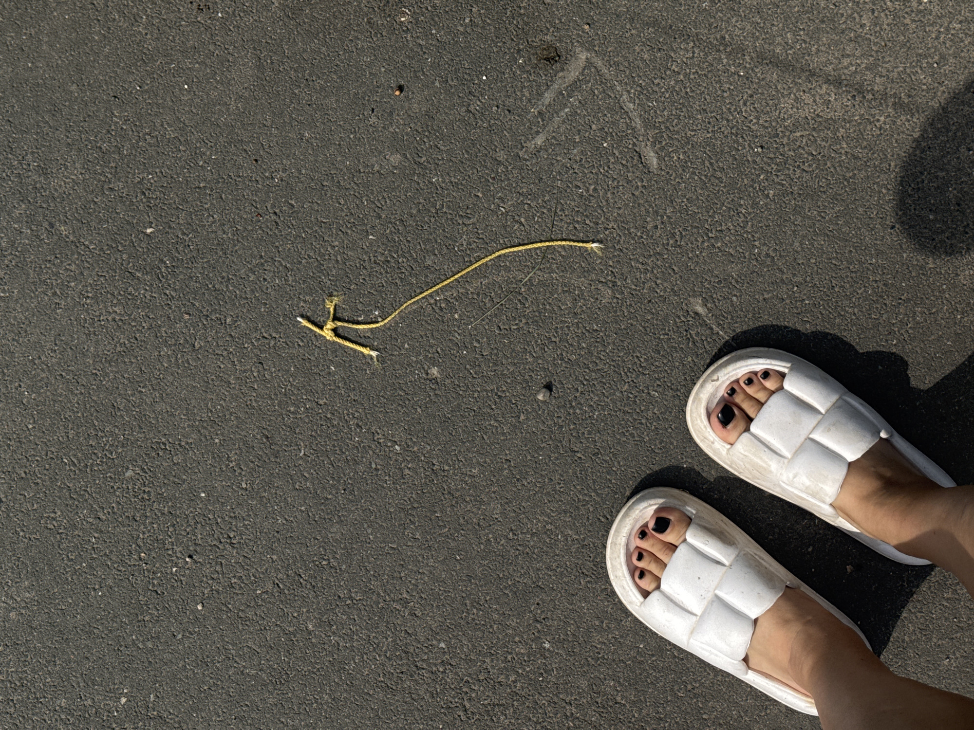 A top-down, eye-level shot shows two bare feet wearing white slide sandals with a woven pattern, standing on dark asphalt. The toes, painted with black nail polish, are visible. To the right and slightly above the feet, a piece of yellow twine is knotted and trails downwards, with a single strand extending further down. The asphalt surface is textured and appears to be a road or sidewalk. The lighting suggests it is daytime, casting subtle shadows. The scene is set in the city of Tolyatti, Russia.