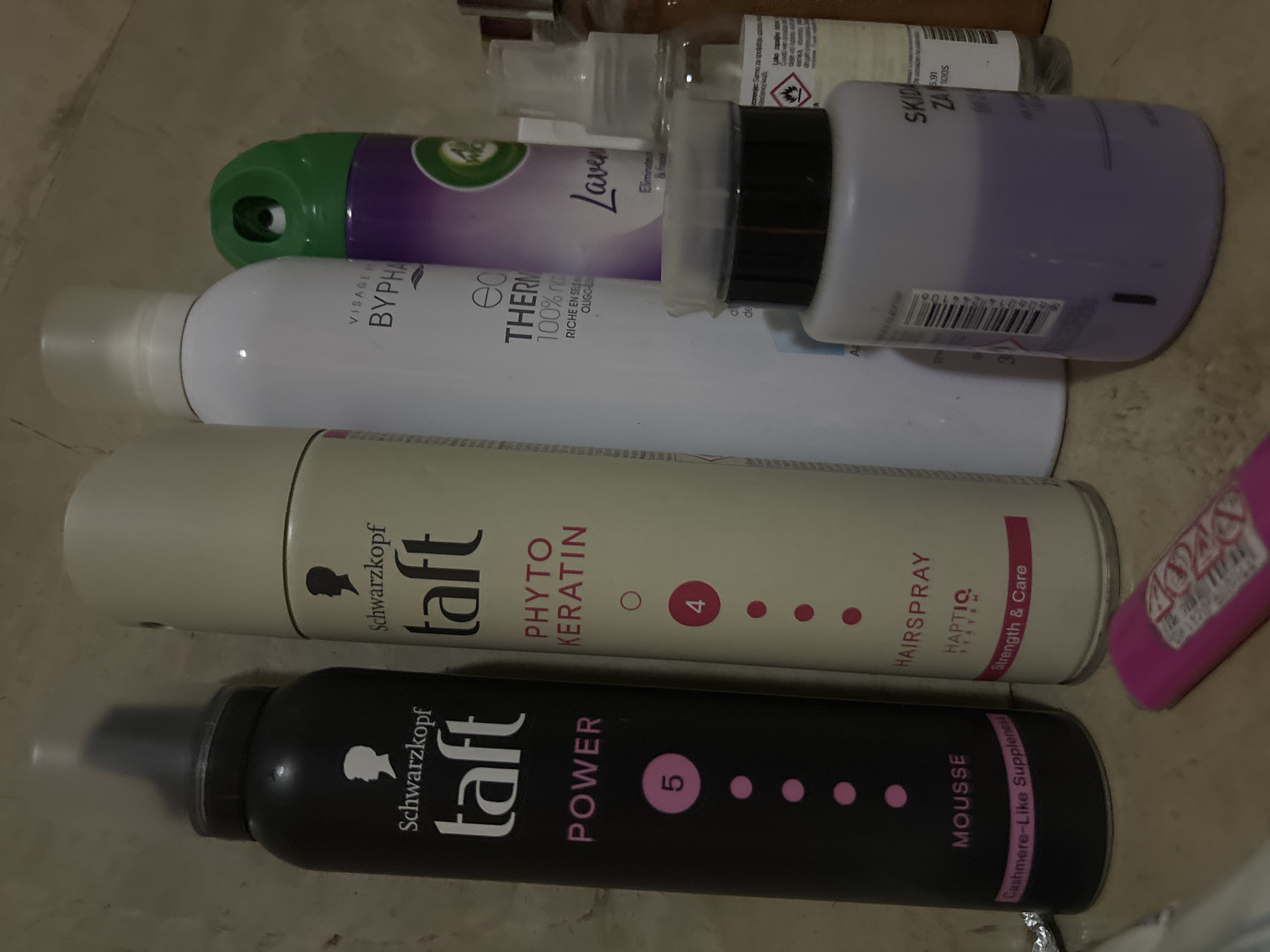 The image shows a collection of personal care products on a flat surface, likely a floor. There are two cans of Schwarzkopf taft hair products: a black can labeled "POWER" MOUSSE and a white can labeled "PHYTO KERATIN" HAIRSPRAY. Above these are a white can of "VISAGEE BYPHA" thermal product and an Air Wick Lavender refill can in green and purple. A bottle of "SKIDAJ ZA" nail polish remover is also visible. The OCR text extracted from the image includes brand names, product names, ingredients, and warnings in both English and Serbian/Croatian. The image does not contain any information about the city of Budva, Montenegro.