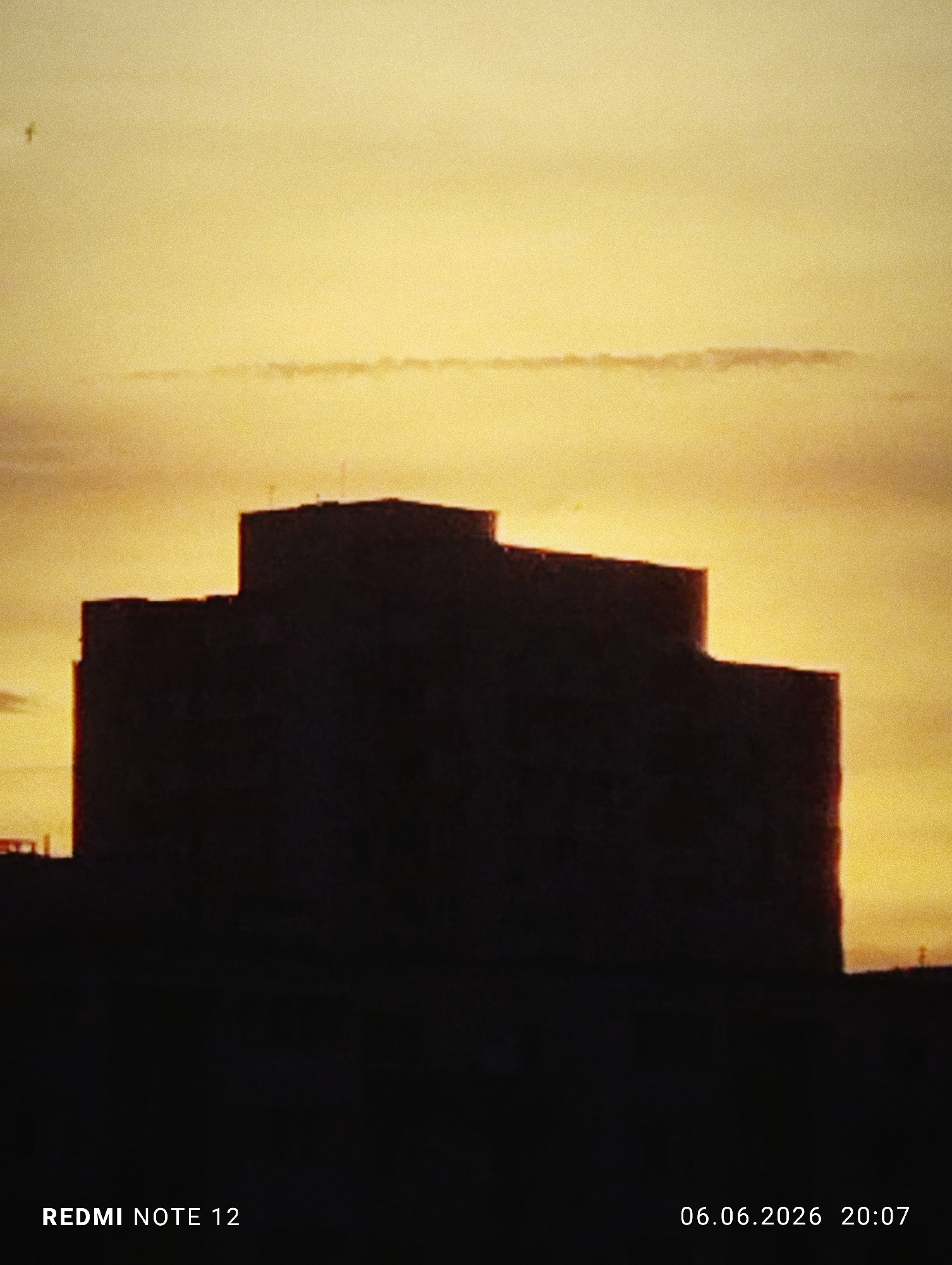 The image depicts an outdoor scene at dusk in the city of Burgas, Bulgaria. The sky, occupying the upper two-thirds of the frame, presents a gradient of warm colors, ranging from a hazy golden-orange near the horizon to a slightly lighter yellow in the upper portions. A distinct, narrow, horizontal cloud band with a subtle reddish tint is visible across the mid-section of the sky. In the upper left quadrant, a small, dark, indistinct form of a bird is captured in flight against the illuminated sky.
Dominating the lower and central portions of the image is the dark silhouette of a large, multi-tiered building. The building exhibits a blocky, stepped architectural profile with varying heights and sections, creating a strong, hard-edged outline against the bright sky. Faint details, possibly an antenna or a small cross, are discernible on the highest visible rooftop section towards the center-right of the building's silhouette. Due to strong backlighting, the building's internal features are completely obscured, rendering it as a solid, dark mass. The immediate foreground below the building is also in deep shadow, appearing as an undifferentiated dark area, concealing any ground-level details or structures.
No individuals or other discernible subjects demonstrating action or interaction are present within the frame.
Two text overlays are present: "REDMI NOTE 12" is located in the lower-left corner, identifying the device used to capture the image. "06.06.2026 20:07" is located in the lower-right corner, indicating the date as June 6, 2026, and the time as 8:07 PM. The overall image displays a noticeable level of grain or noise, consistent with digital capture in low-light conditions.