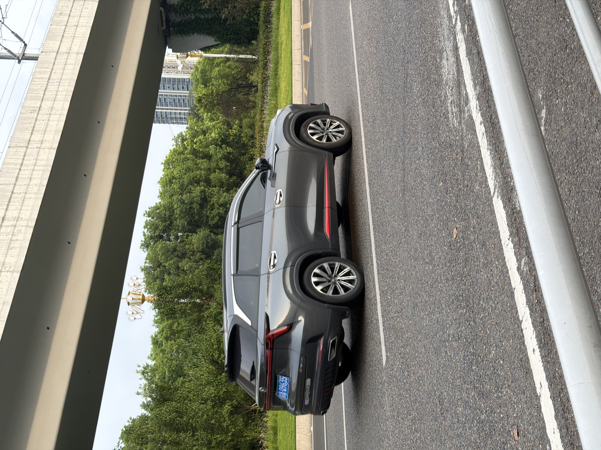 A dark gray SUV, identified by the license plate "A 09635," is driving on a paved road in the city of Jiangjin, China. The vehicle is positioned in the right lane of the road. The surrounding environment consists of a lush green area with trees and grass to the left of the road, and an elevated structure, possibly a bridge or overpass, spanning overhead. Buildings and a lamppost are visible in the background, suggesting an urban setting. The road has clear white lane markings. The scene appears to be captured during daylight hours.
