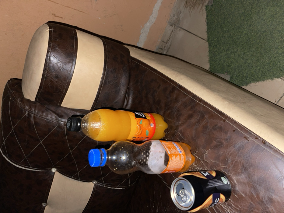 A leather sofa with brown and beige cushions is partially visible. The sofa's arm is adorned with bottles and a can. A partially full orange juice bottle sits near the top of the armrest, below that is a nearly empty soda bottle with a blue cap. A black and gold can lies near the edge. The background reveals a wall, tiles, and artificial green grass.