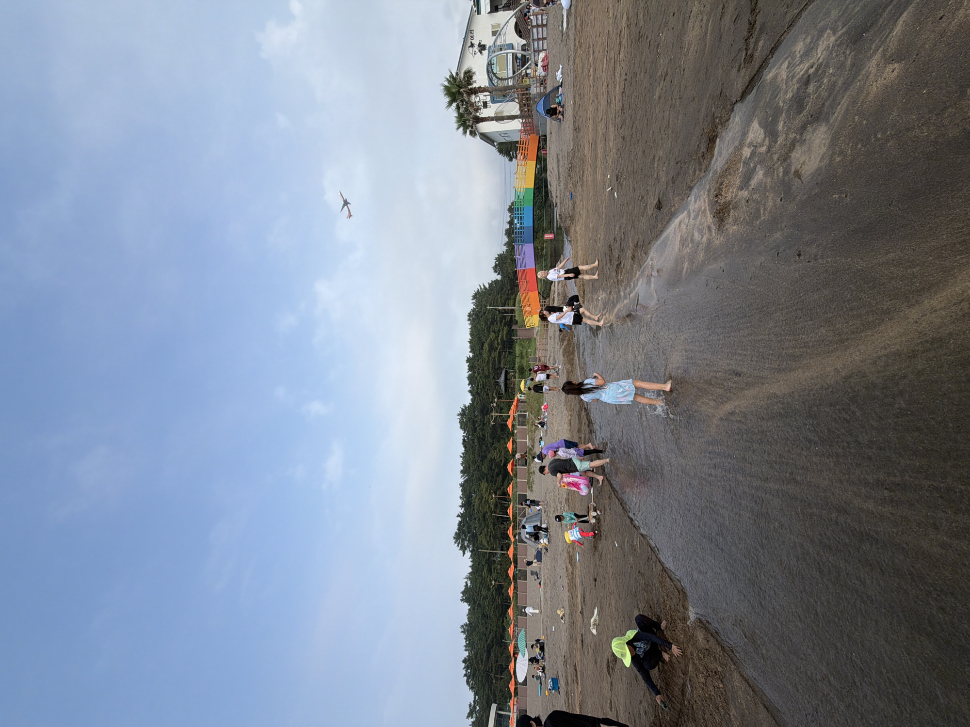 An aerial shot captures an orange and white airplane flying in a clear blue sky over a sandy beach in Jeju, South Korea. On the beach, various people are scattered, some in the water and others on the sand. A group of children are playing in a shallow stream that runs across the sand. In the background, rows of orange umbrellas provide shade for beachgoers, and a colorful rainbow bridge spans over the stream. A building with palm trees in front of it sits further down the beach.