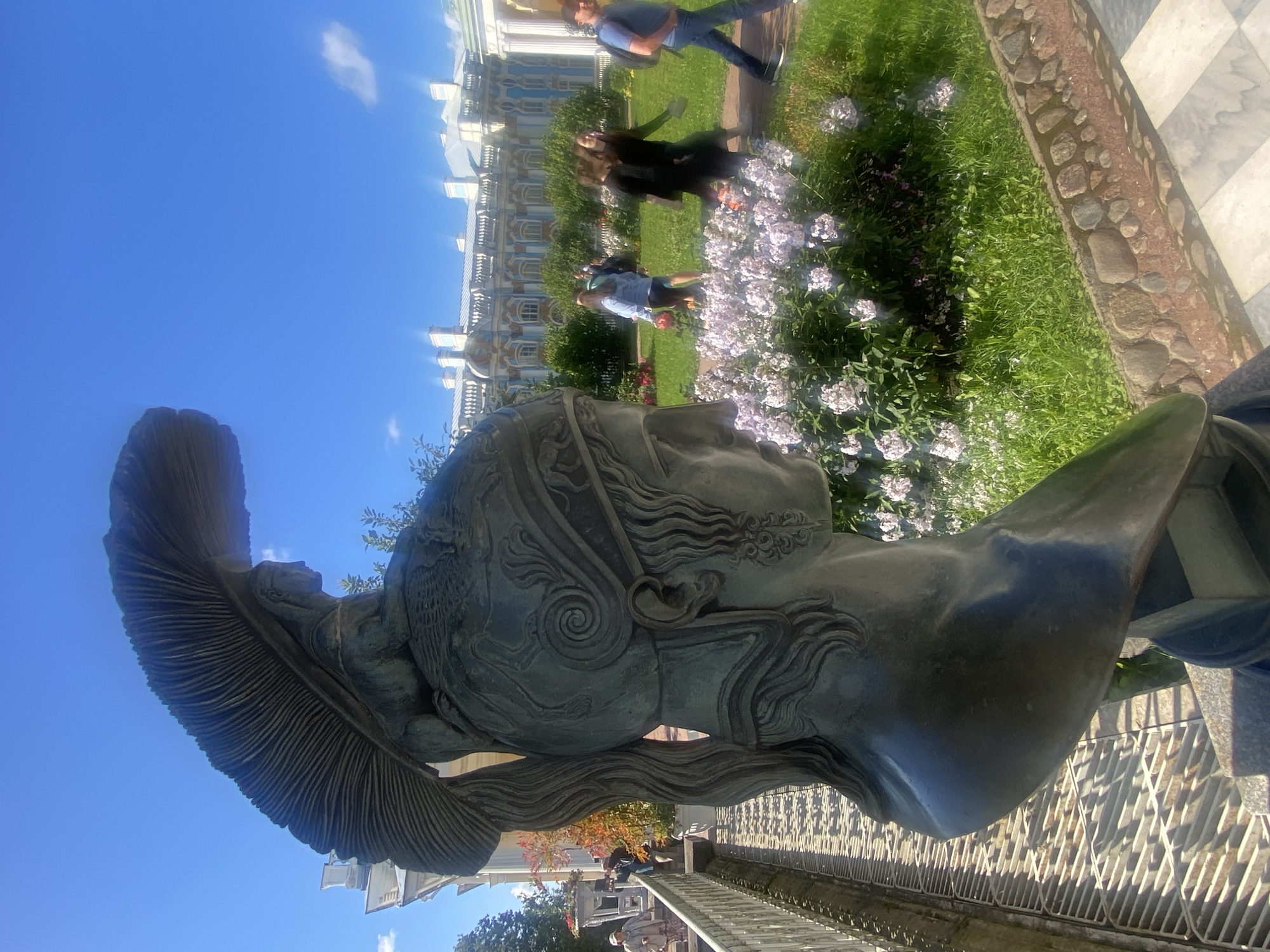 A bronze bust of a Roman soldier wearing a plumed helmet is in the foreground, facing right, against a bright blue sky. Behind the bust, a group of people are walking on a cobblestone path next to a lush green lawn dotted with pink and white flowers. In the background, a grand blue and white palace with many windows and ornate architectural details stretches across the horizon. The scene is illuminated by bright sunlight, casting shadows on the ground. The people are casually dressed and appear to be tourists. The bust is weathered and shows signs of age. This is likely the Catherine Palace in Pushkin, a suburb of Saint Petersburg, Russia, known for its opulent architecture and gardens.