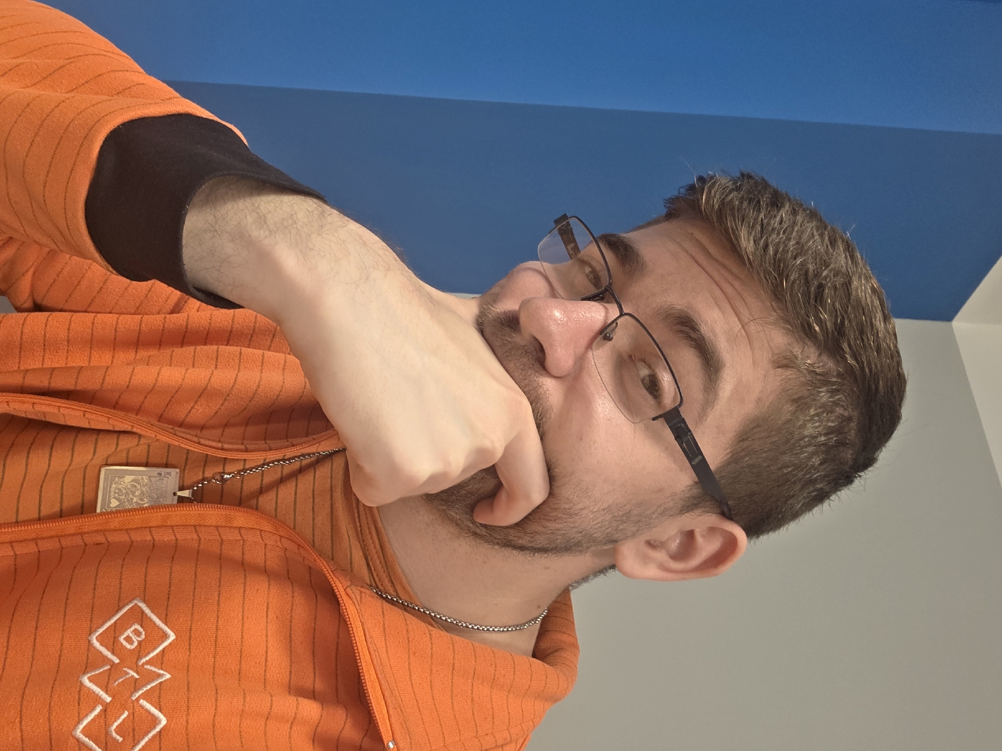 A person with fair skin, short brown hair, and a beard is wearing glasses and an orange zip-up jacket with a white emblem on the left side. The jacket has a black cuff on the sleeve. The person has their hand up to their face, appearing to be touching their cheek with a clenched fist. They are wearing a silver chain necklace, and another pendant is partially visible inside the jacket. The background consists of a blue ceiling meeting a white wall. There's no information to determine if the location is the city of Plovdiv, Bulgaria.