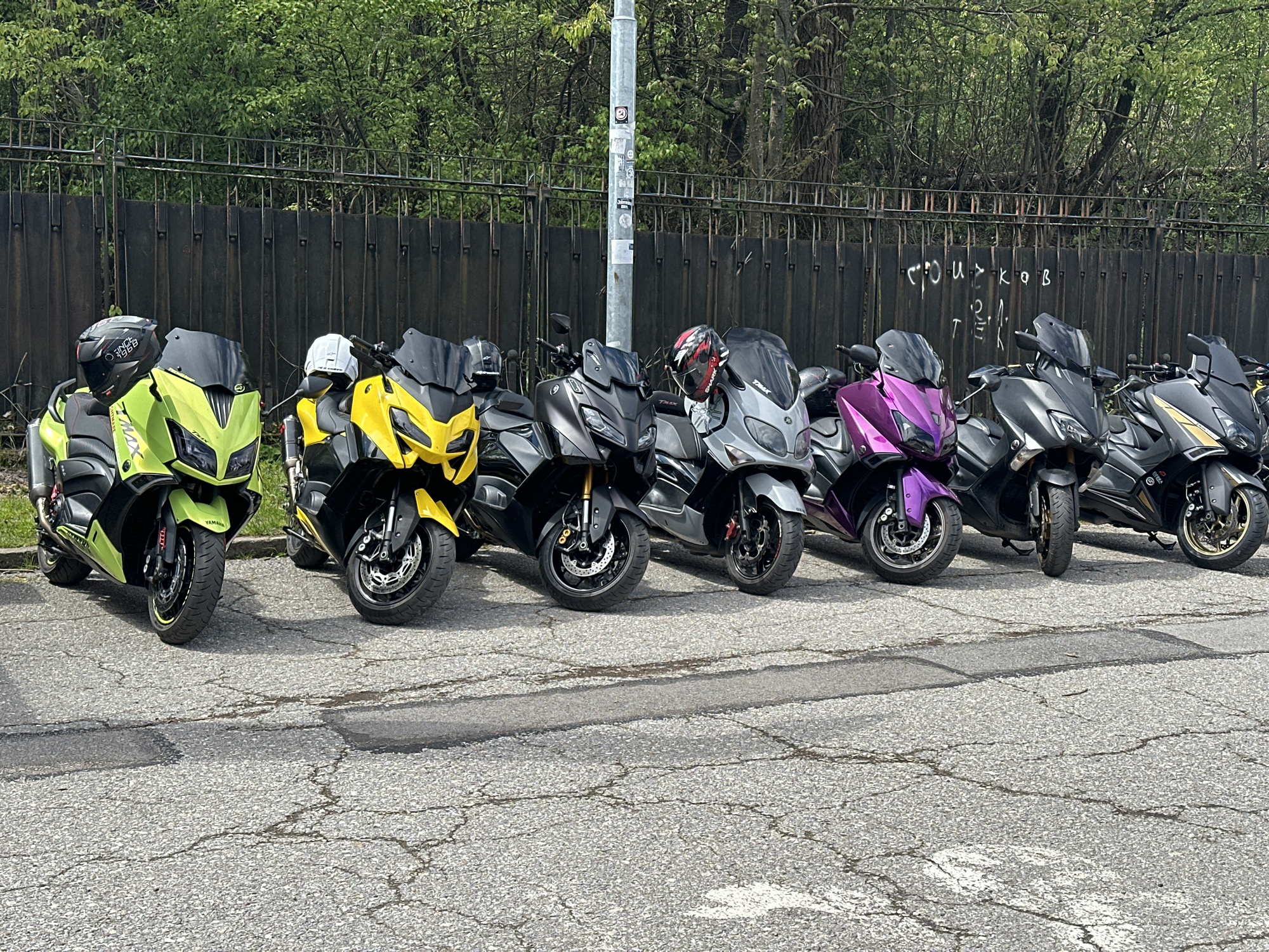 The image shows a row of seven parked Yamaha TMAX scooters in Sofia, Bulgaria. The scooters are parked on an asphalt road with visible cracks and patches. The bikes are colored green, yellow, black, grey, purple, and black with gold trim. A metal fence runs along the background, with green foliage and trees visible behind it. A light pole stands beside the third motorcycle from the left. Graffiti is visible on the fence behind the scooters.