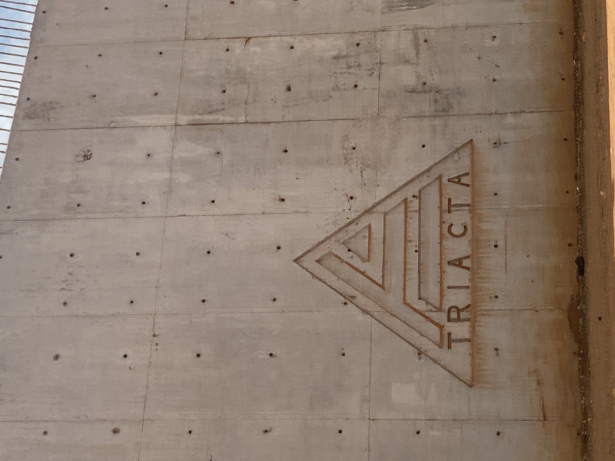 The image shows a close-up, eye-level, full shot of a concrete wall with the word "TRIACTA" carved into it, centered below a triangular logo. The wall is made of large, rectangular concrete slabs, showing vertical and horizontal seams. Numerous small, dark holes are scattered across the concrete surface, consistent with construction materials or imperfections. The word "TRIACTA" is etched in a rough, industrial font. The logo above it is an abstract triangle with internal lines creating a layered or maze-like effect. The bottom portion of the wall shows streaks of reddish-brown discoloration, possibly from rust or water runoff, and a small pile of dark debris is visible at the base of the wall. The ground in front of the wall is sandy and dusty. The overall scene appears to be an outdoor construction site or a utilitarian structure in Nyanya, Nigeria.