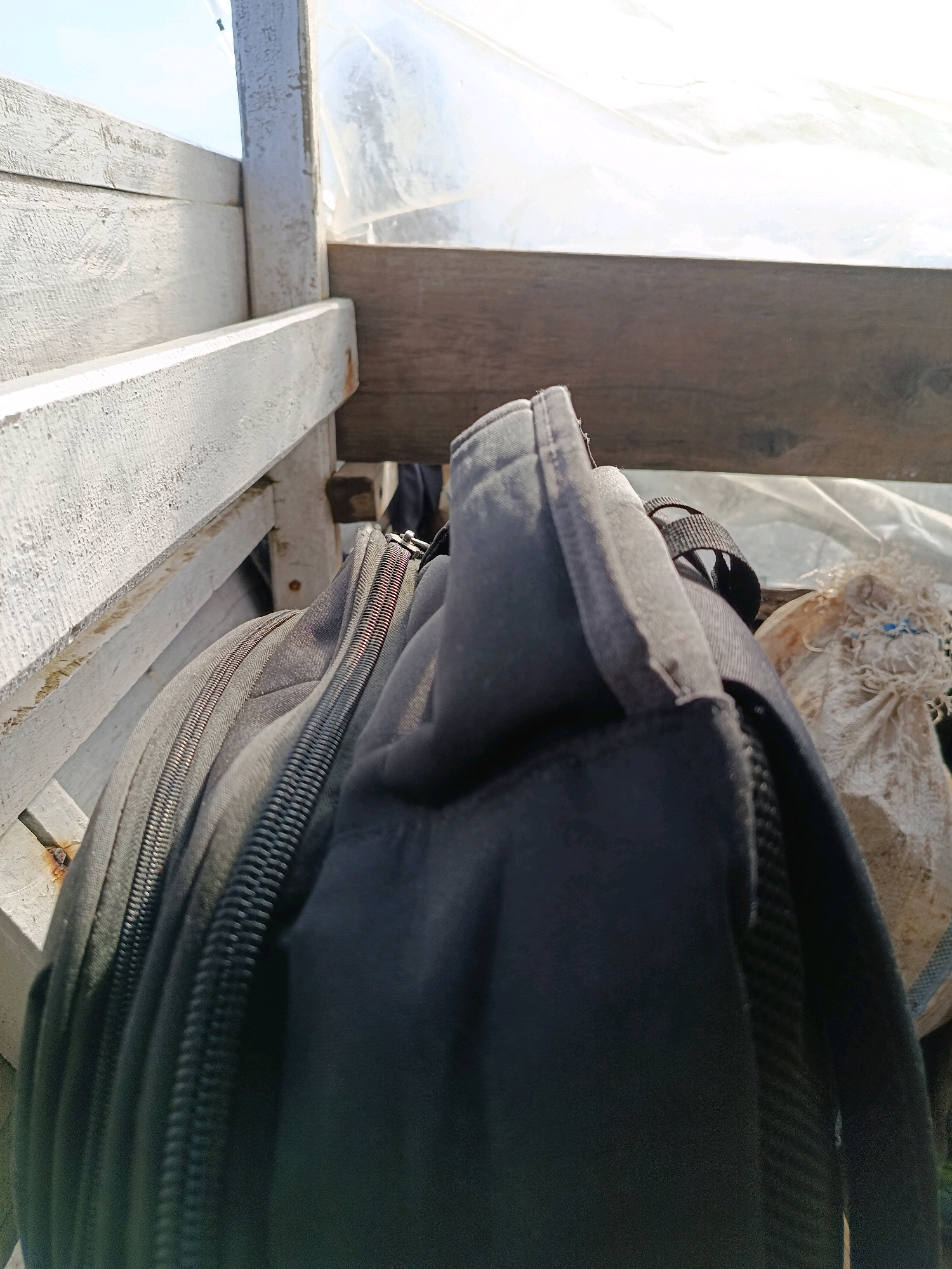A close-up shot focuses on a black backpack and a white wooden structure. The backpack's zipper is prominently featured, with its teeth and pull tab visible. To the right of the backpack, a burlap sack is partially visible, filled with what appears to be dried vegetation. The background reveals a translucent plastic sheeting, possibly from a greenhouse or makeshift shelter, illuminated by bright sunlight. The wooden structure, painted white and weathered, forms the left and top borders of the frame, suggesting an outdoor setting. The scene is likely captured in Gong-Gong, Indonesia, given the context provided.