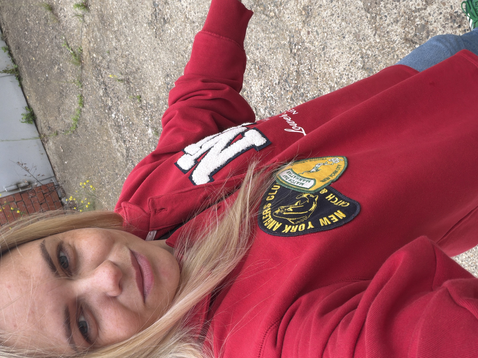 A young Caucasian woman with blonde hair and blue eyes is taking a selfie outdoors. She is wearing a red sweatshirt with patches and a large white "N" with a blue outline on the front. The background appears to be a concrete area with some scattered vegetation and a grey building. The image does not contain any specific indicators to determine the location within Votkinsk, Russia.
