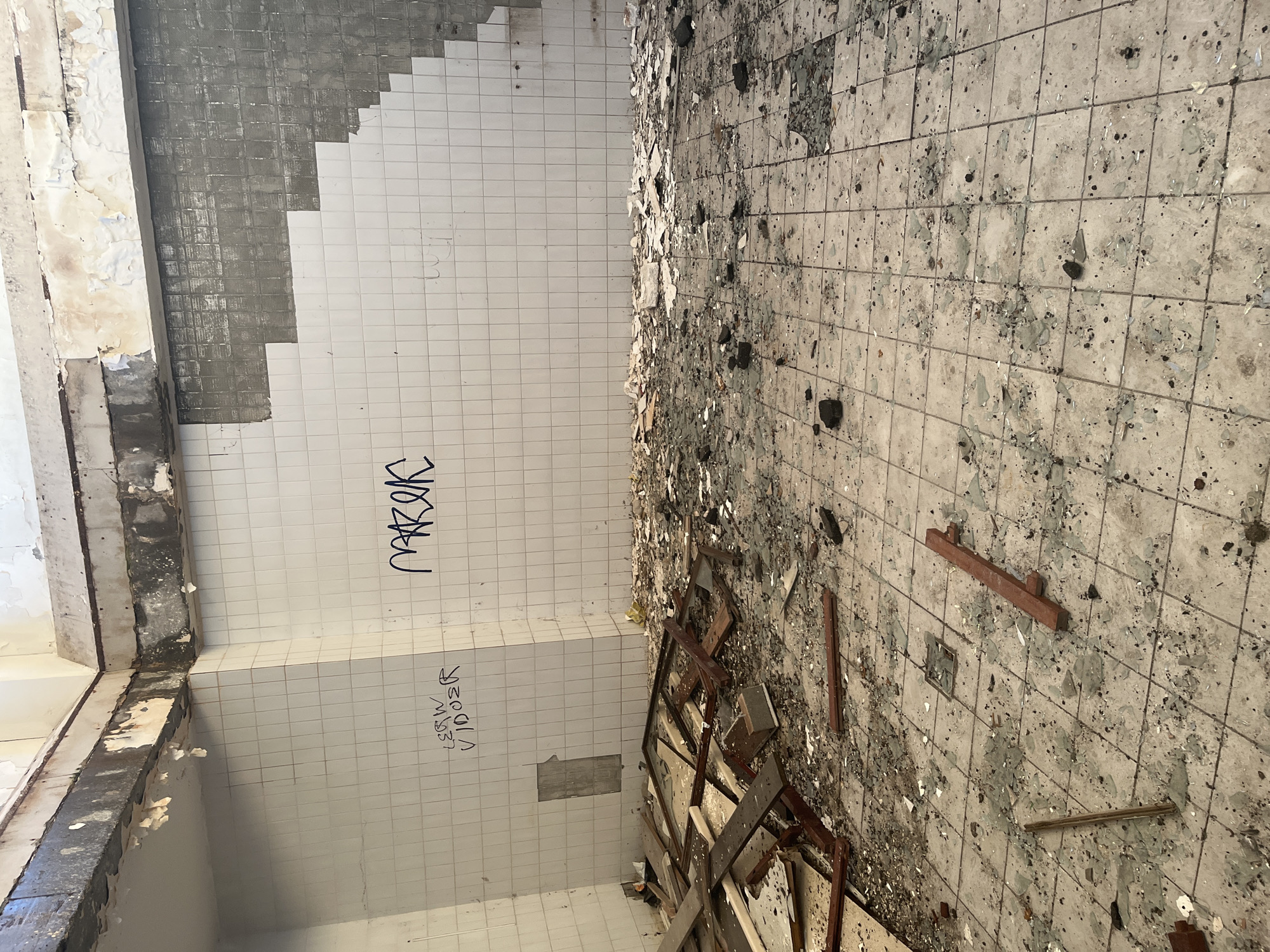 The image depicts the interior of a derelict building in what appears to be Марковский, Russia. The room is heavily damaged, with debris scattered across the floor and walls. The walls are tiled with white tiles, some of which are missing or damaged. Graffiti is visible on the walls. The floor is also tiled, with many tiles broken or missing. There is wooden debris and other unidentified objects strewn around the floor. The overall impression is one of abandonment and decay. There are no visible people.