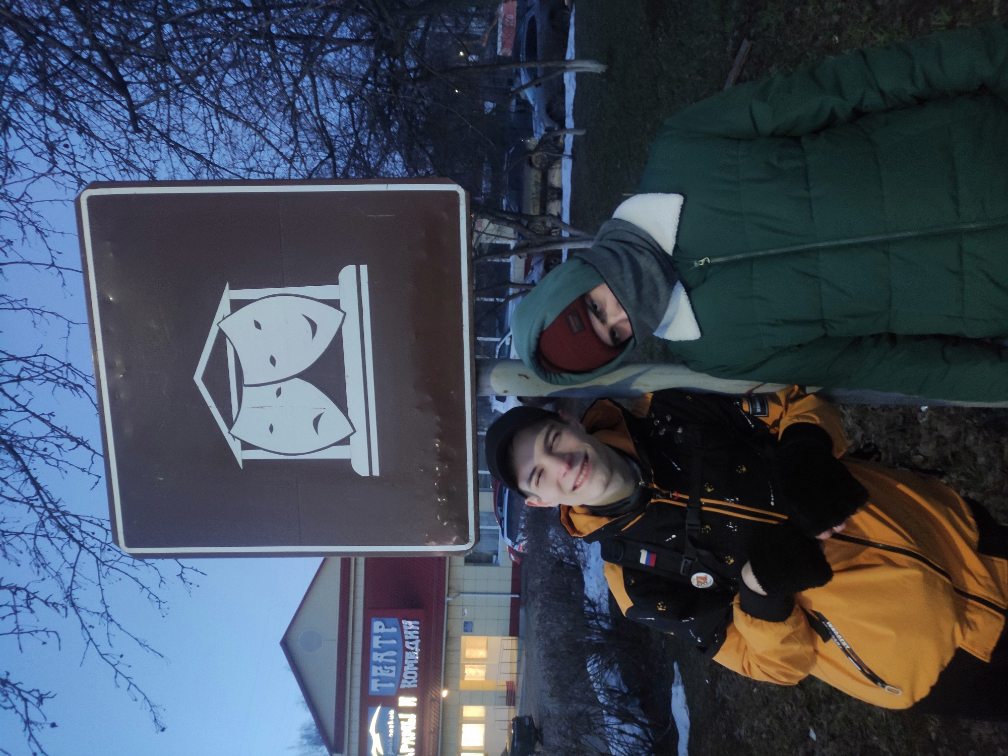 Two people are posing near a brown sign with white theatrical masks. The person in the foreground wears a black and yellow jacket, a black cap, and black mittens. The other person is wearing a green jacket with a grey hood and a red hat underneath the hood. Behind them is a building with a sign that reads "ТЕАТР АРТИСТЫ И КОМЕДИИ" which translates to "THEATER ACTORS AND COMEDIES." The scene appears to be outdoors, possibly in Chaikovsky, Russia, given the Russian signage. Bare tree branches are visible in the background.