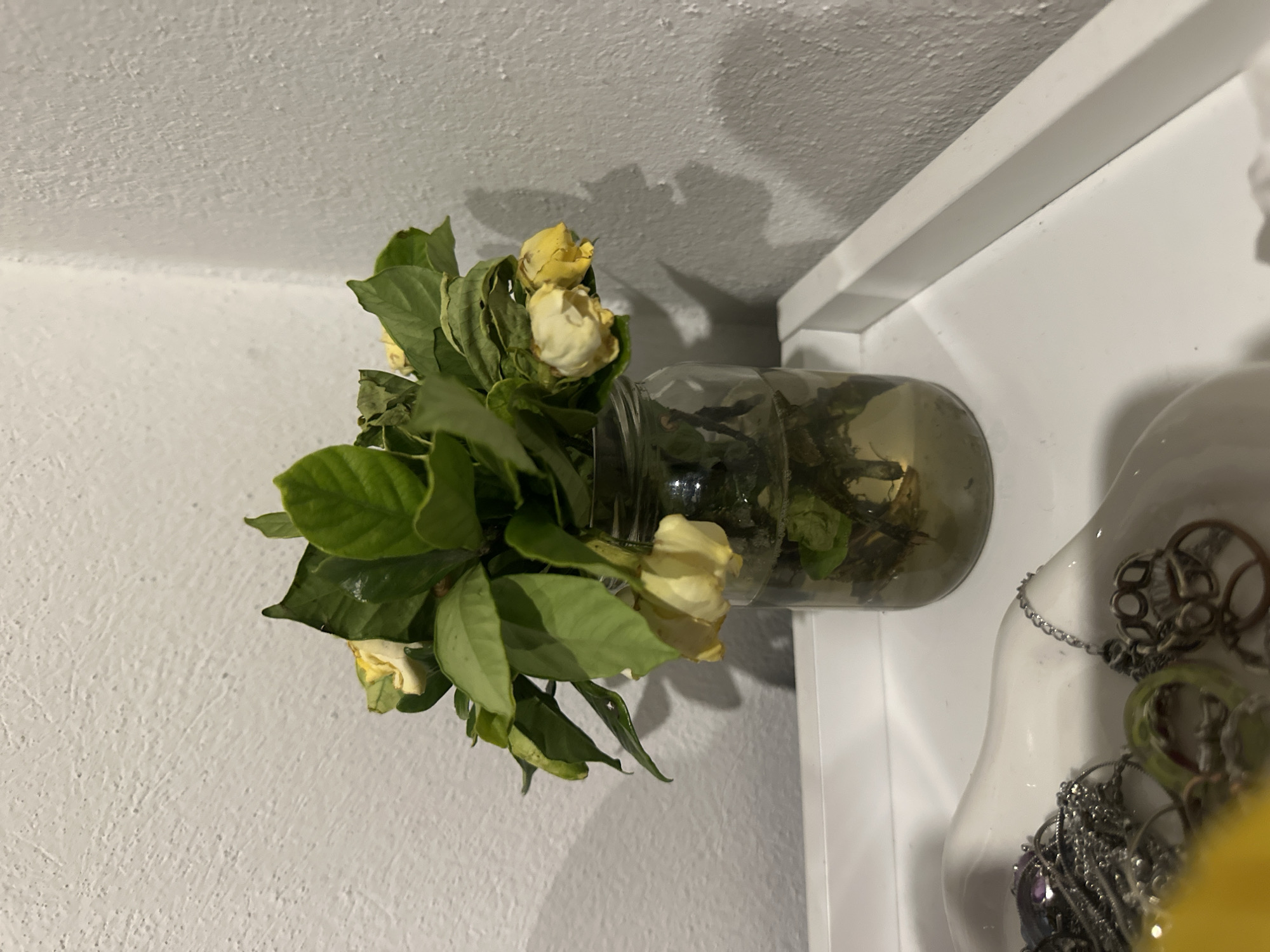 A bouquet of yellow roses with green leaves sits in a clear glass jar filled with murky water. The bouquet is on a white shelf next to a white mannequin bust adorned with various necklaces. The background consists of a textured white wall and a white ceiling. The setting appears to be indoors, possibly a home or retail space. The city of Huixquilucan de Degollado, Mexico, is not directly discernible from the image's content.