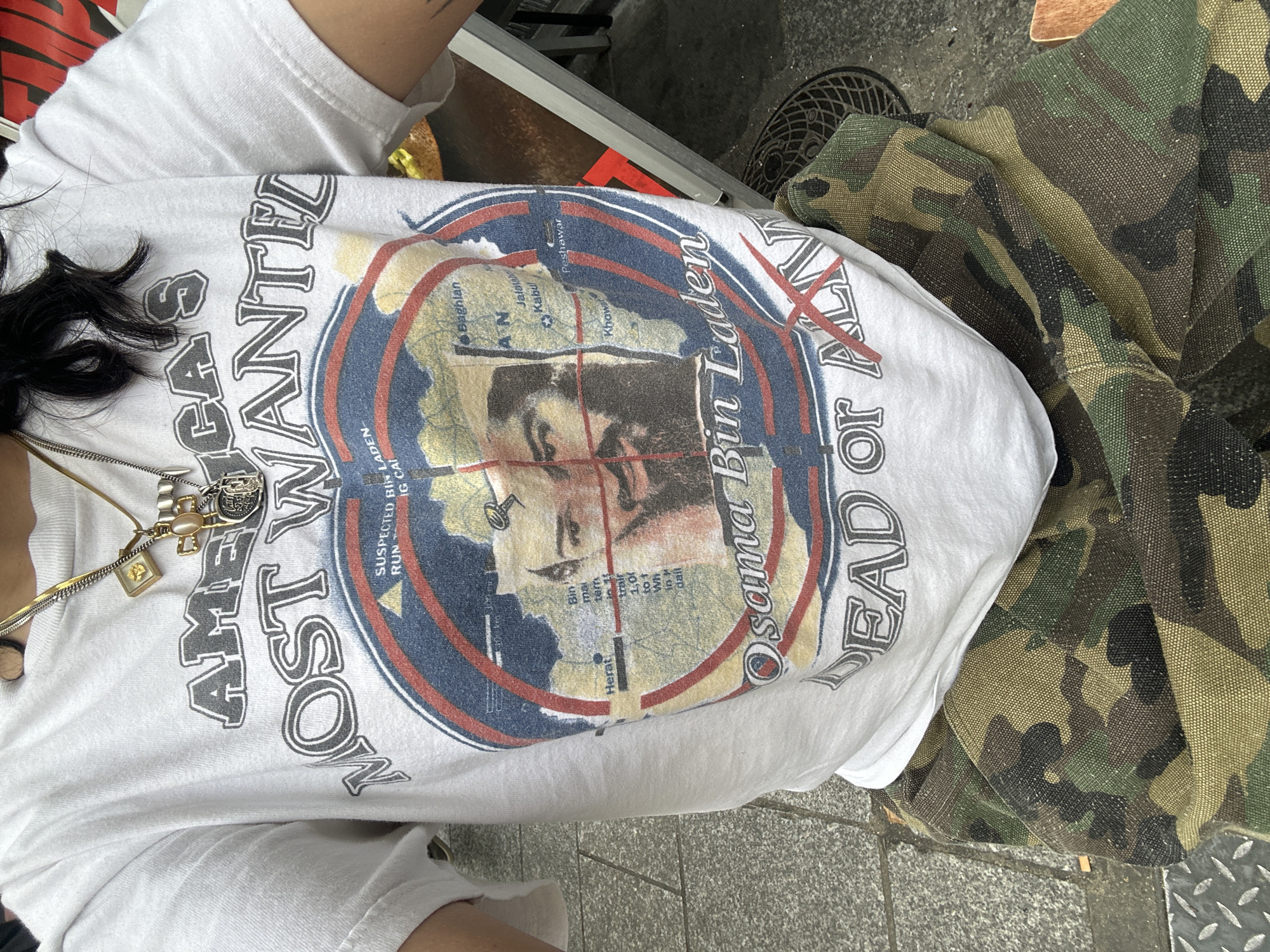 A person wears a white t-shirt featuring an image of Osama Bin Laden. The shirt reads "America's Most Wanted, Osama Bin Laden, Dead or Alive," surrounding the image. The person is also wearing camouflage pants. The location is possibly Paris, France, due to the presence of the "IG CAI" text on the t-shirt, which indicates an abbreviation for "Inspection Générale des Carrières" which is related to the Catacombs of Paris. The setting appears to be an urban environment, with a sidewalk and possibly a sewer grate visible.