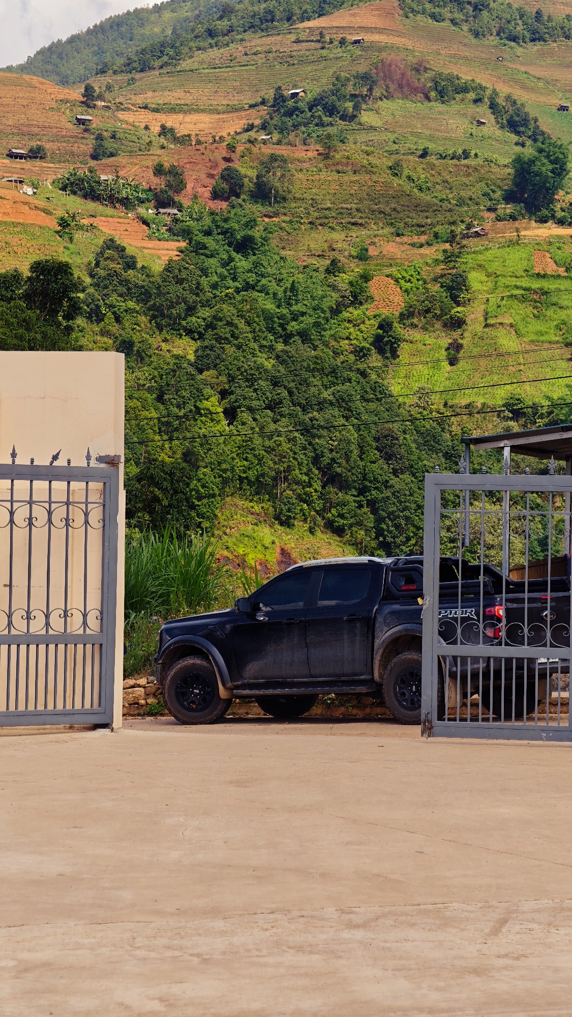The vertical image presents a vibrant rural landscape, likely in Lao Chai, Vietnam, dominated by a steep, terraced hillside. In the foreground, a dark-colored, likely black or dark grey, Ford Ranger Raptor pickup truck is parked on a light-colored paved surface, partially obscured by two open metal gates. The gates are painted a light grey and feature ornate, symmetrical scrollwork. To the immediate left of the left gate, a section of a light beige building wall is visible.
Behind the truck and gates, the landscape unfolds into a dramatic mountain slope. This hillside is characterized by extensive terraced fields, displaying a mosaic of rich green vegetation and reddish-brown cultivated earth. Patches of dense green forest are interspersed among the agricultural areas. Numerous small, simple huts or houses are dotted across the distant slopes, blending into the environment. Faint electrical wires can be seen stretching across the midground.
The scene is illuminated by bright, clear daytime sunlight, suggesting a cloudless sky, likely late morning or early afternoon. The truck appears somewhat dusty or muddy, hinting at its use in rugged terrain. The only visible text is "RAPTOR" on the tailgate of the pickup truck. No people are present, and the overall atmosphere is one of serene rural life where modern vehicles coexist with traditional farming practices.